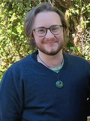 Scout wears glasses and smiles standing in front of an outdoor, sunny, green background