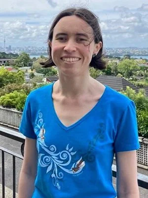 Áine smiles wearing a t-shirt and standing on a balcony with a green cityscape behind.