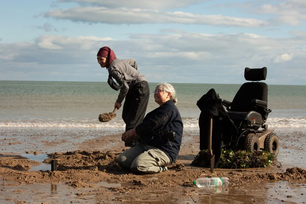 Liz is laughing, sitting close to her power chair, just up from the shoreline at the beach, her hands covered in sand, with another member of the team beside her, also smiling and holding a sand-covered spade.