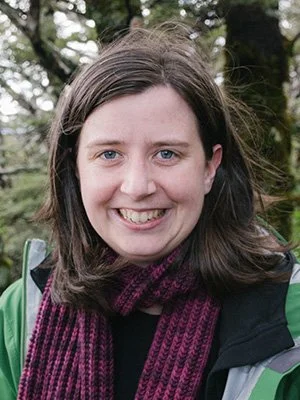 Raven wears a coat and scarf and smiles for the camera in front of out-of-focus trees