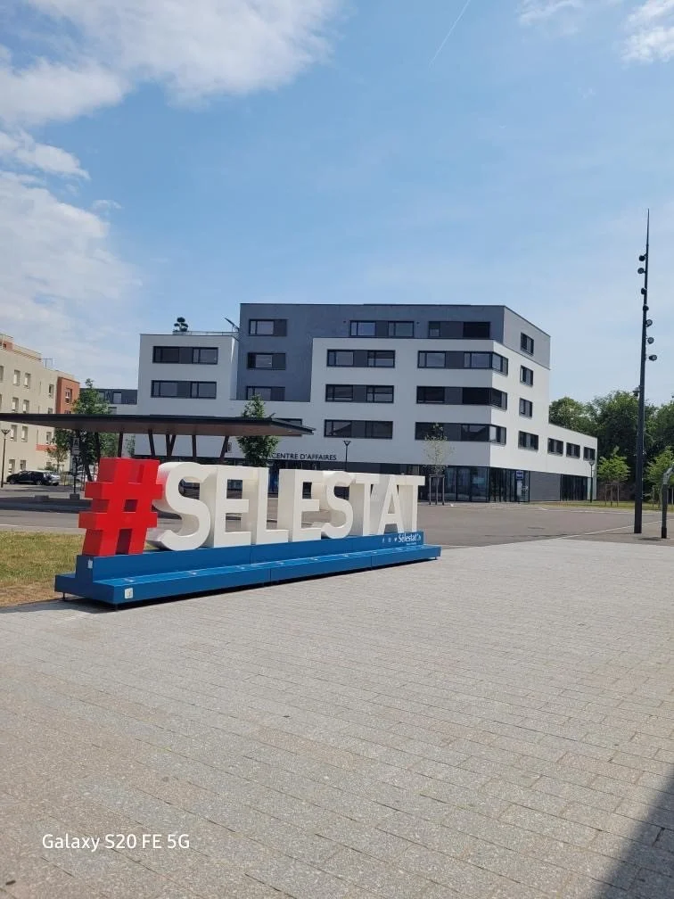Hashtag géant "#SELESTAT" devant un centre d'affaires moderne sous un ciel bleu.