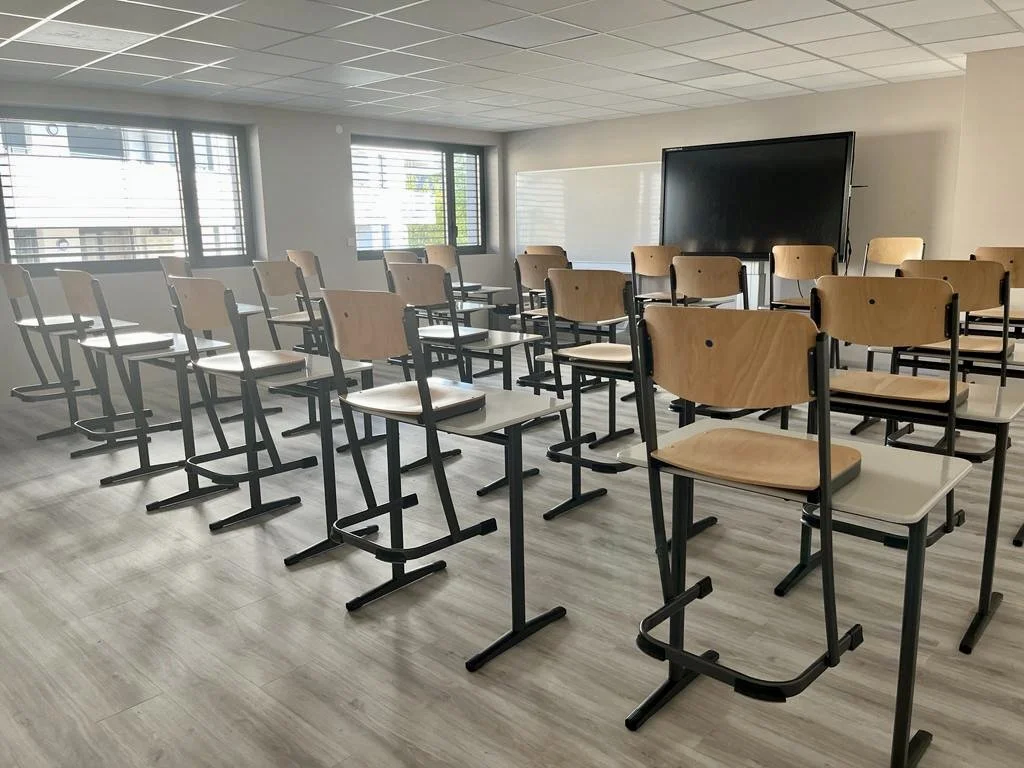 Une salle de classe vide avec des chaises en bois sur les bureaux, un tableau blanc et un écran.