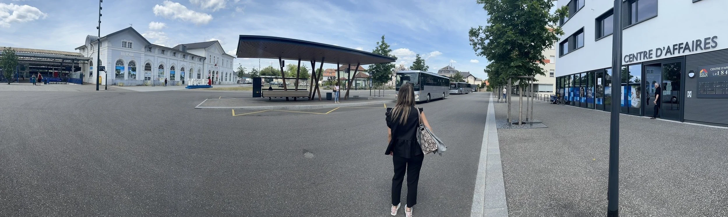 Une personne marche vers une gare routière moderne avec un bâtiment ancien à gauche, un abri d'attente et des bus garés. À droite, un centre d'affaires avec des vitrines et l'inscription 'CENTRE D'AFFAIRES'.