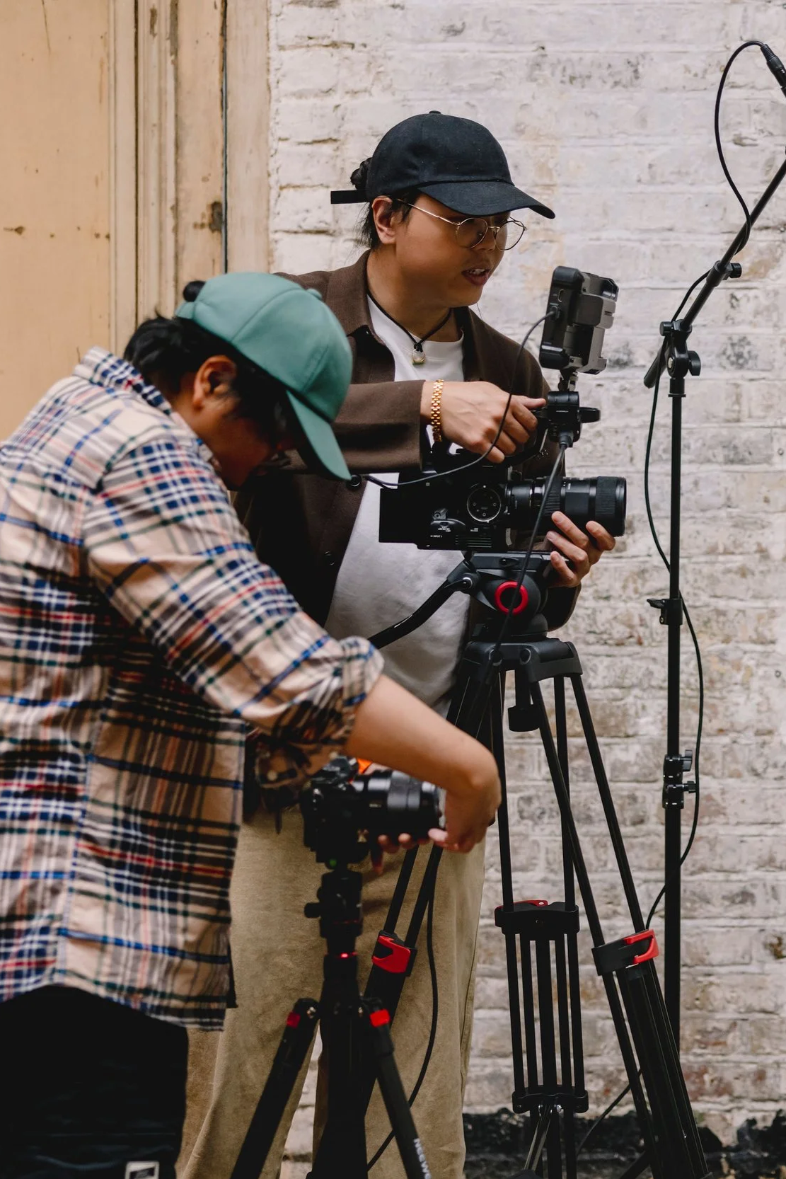 Two people operating camera equipment indoors against a white brick wall, one adjusting the camera on a tripod and the other holding a smaller camera.