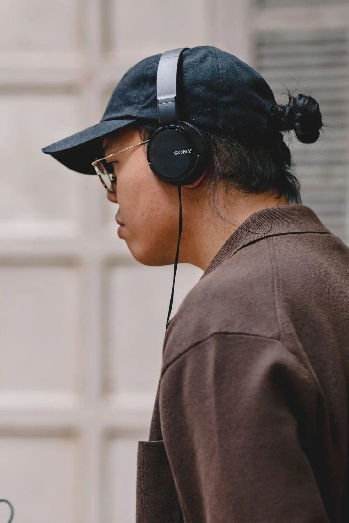 A person wearing a black cap, glasses, and Sony headphones, looking down, with a brown sweatshirt.