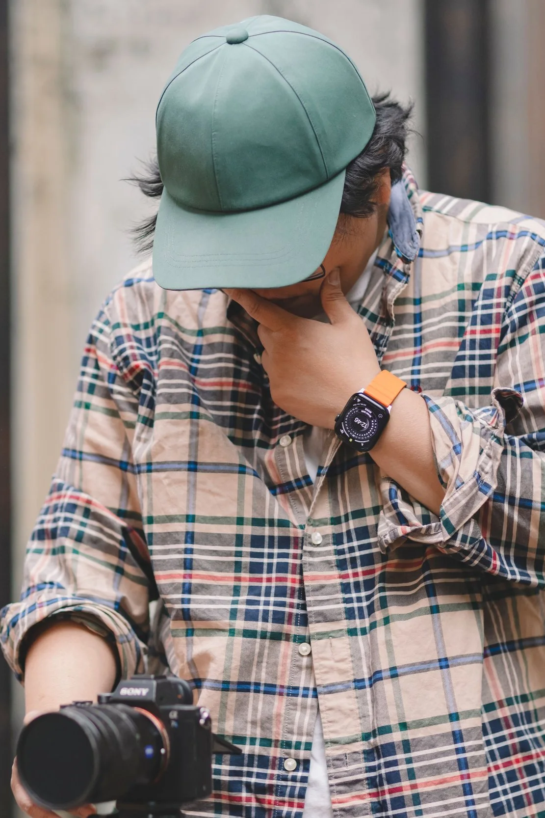 A person wearing a teal baseball cap, plaid shirt, and an orange smartwatch, with their hand on their face, looking down. A camera is positioned in front of them.