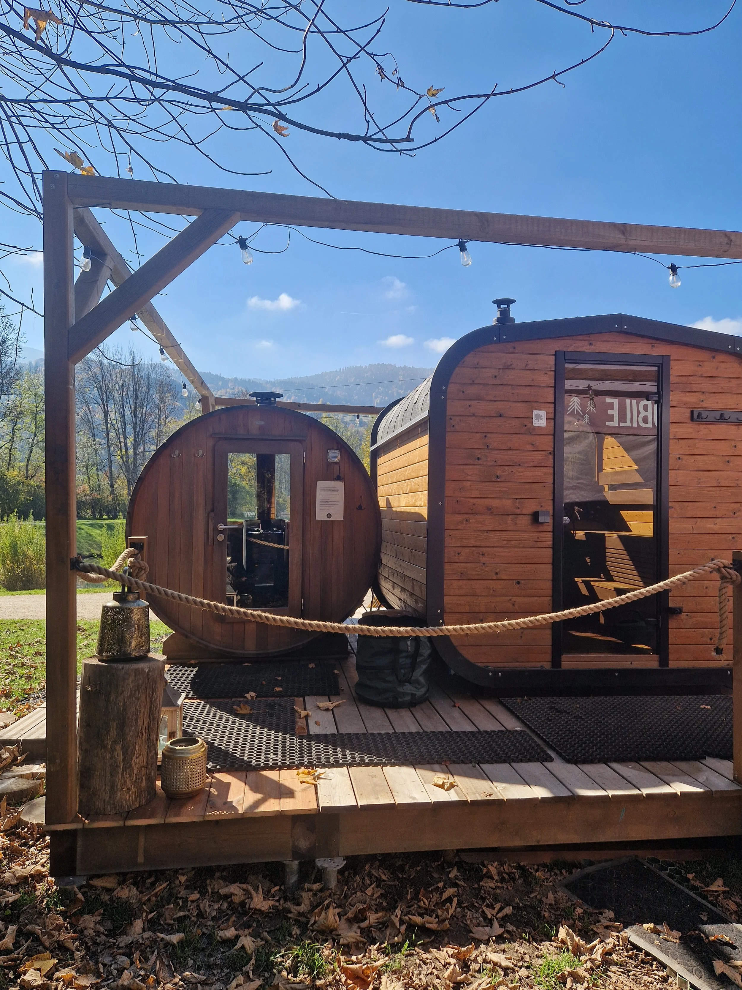 A barrel sauna and a cabin sauna side by side on a wooden platform under a blue sky