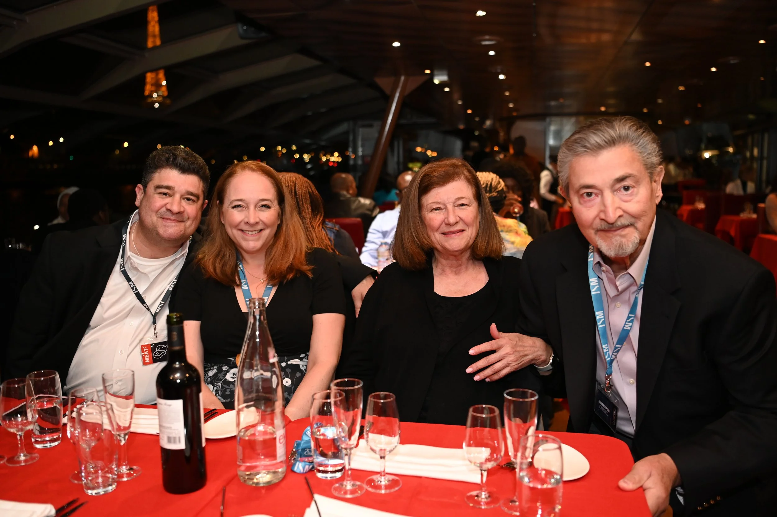 Four people sitting at a table during an evening event in a restaurant or banquet hall, with glasses and bottles on the table, and a cityscape visible through a large window in the background.
