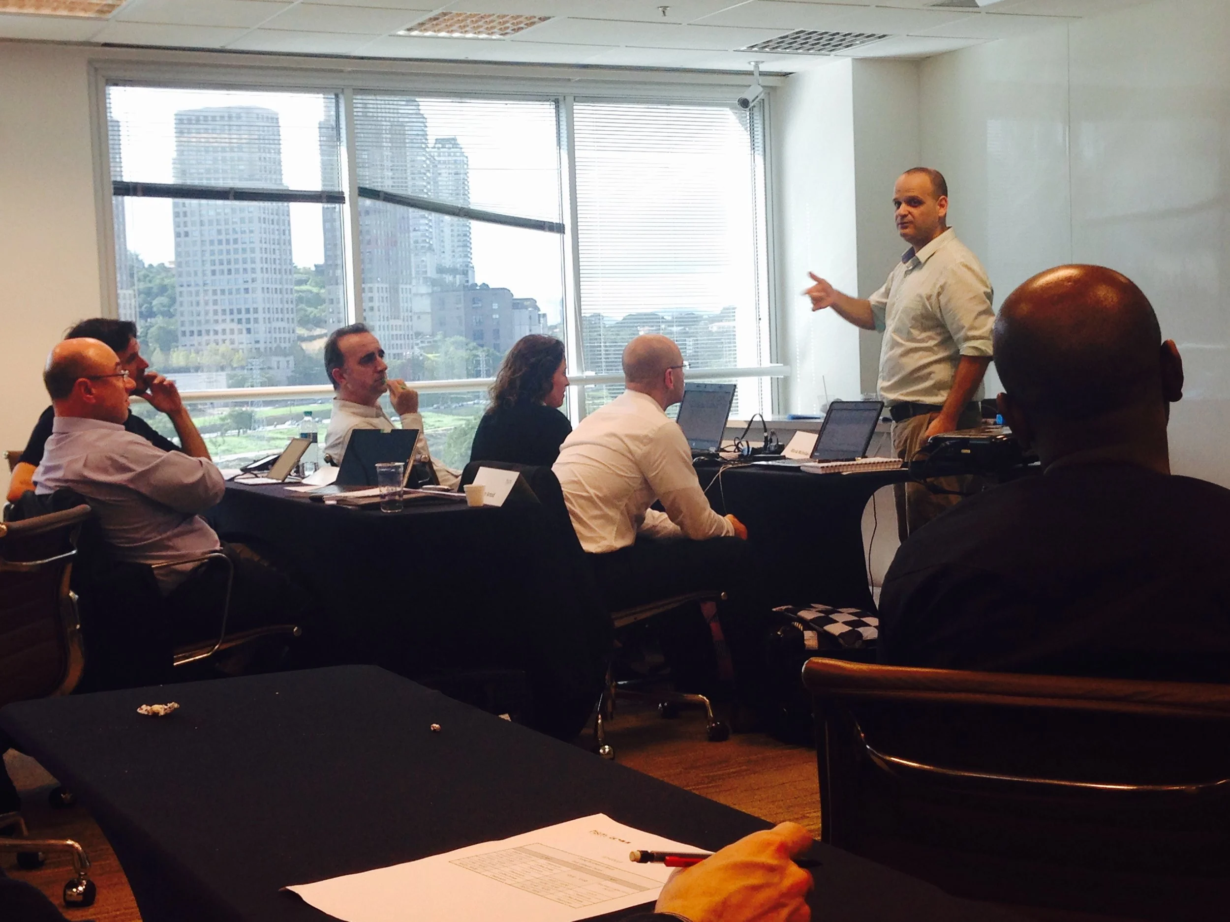 A man stands at the front of a conference room giving a presentation to a group of five people seated at tables. Large windows show a cityscape with tall buildings.