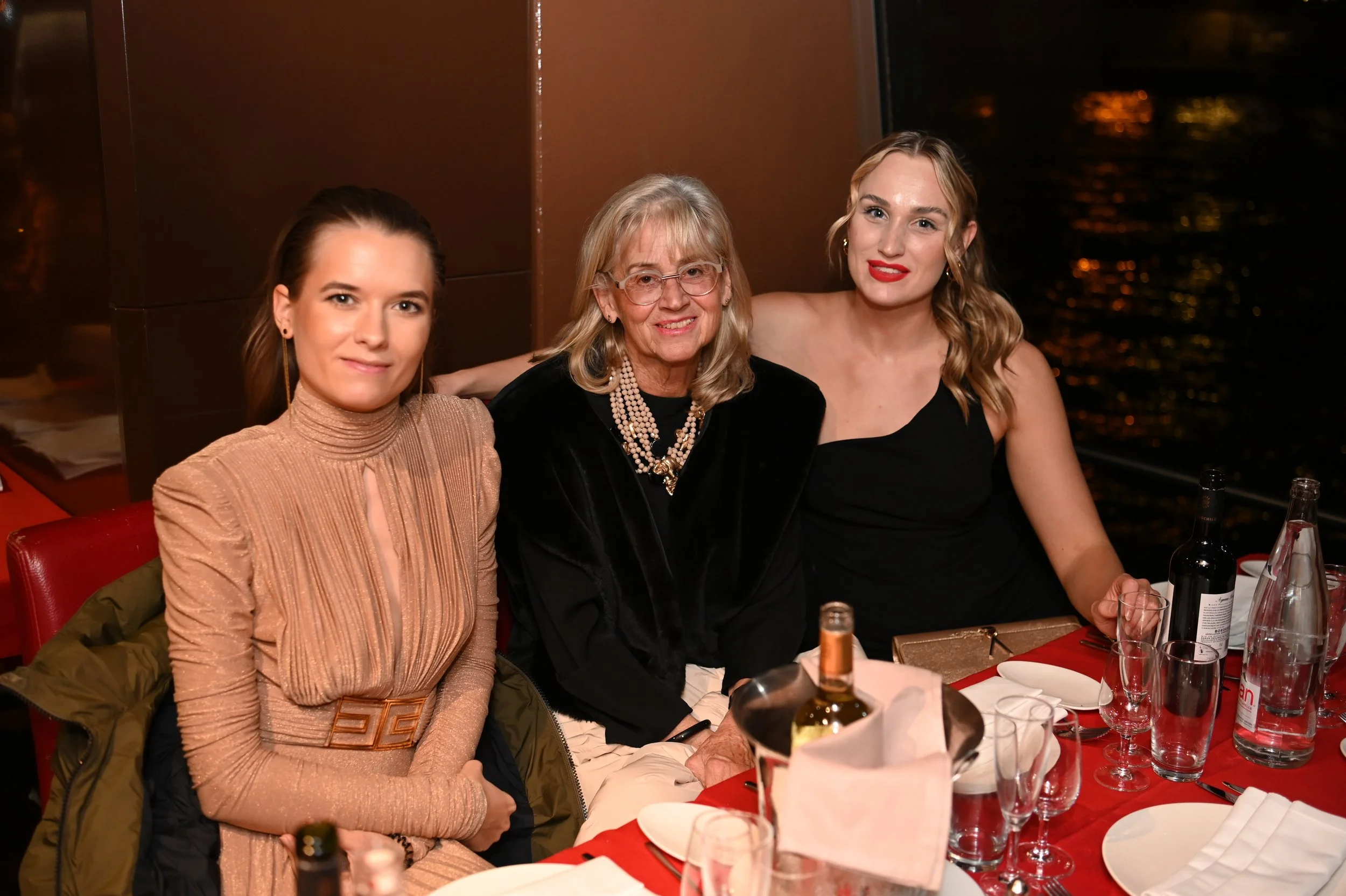 Three women sitting at a dining table during an evening event, with wine bottles and glasses on the table, in front of a window showing a night view.