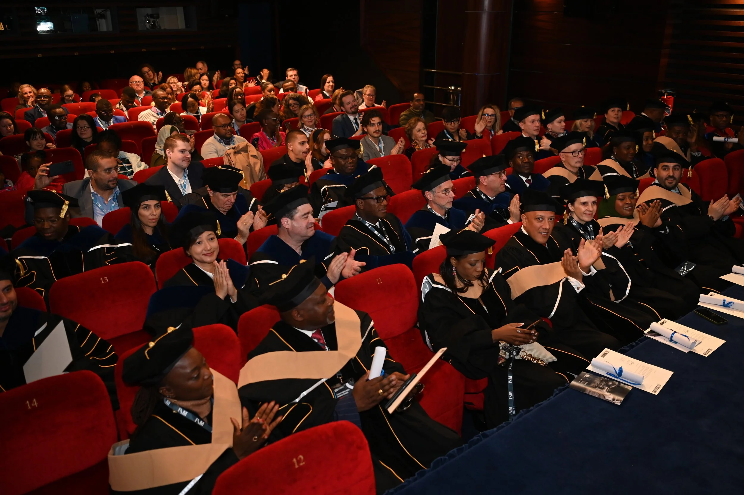 Audience members in academic regalia seated in a theater, clapping during a graduation ceremony.