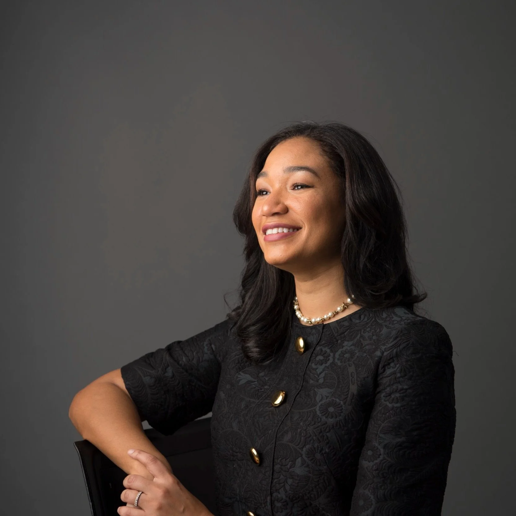 A woman with black hair, wearing a black patterned dress and pearl necklace, smiling and looking to her left against a gray background.