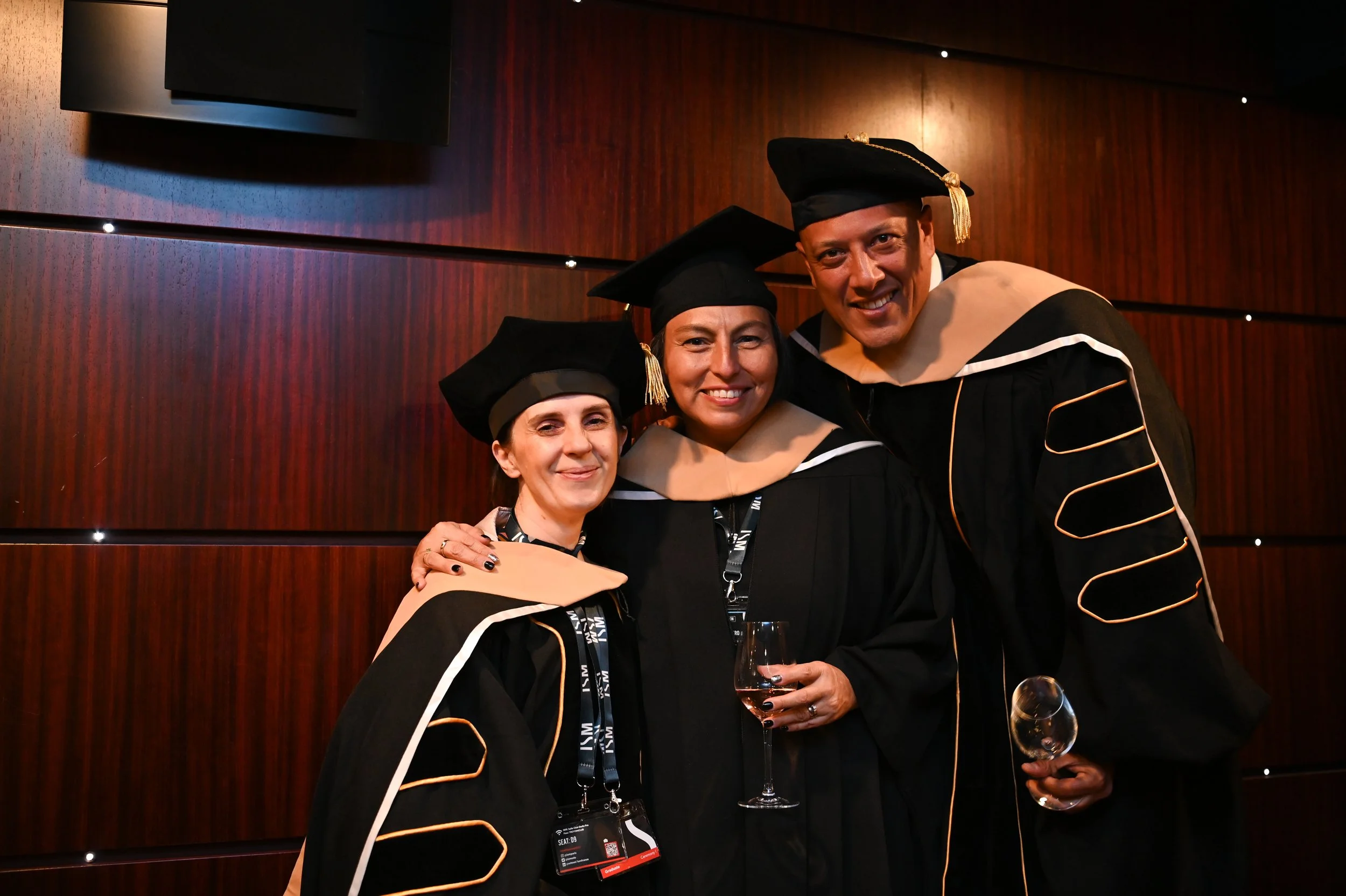 Three graduates in cap and gown smiling at a ceremony, holding wine glasses, standing in front of a wooden wall.