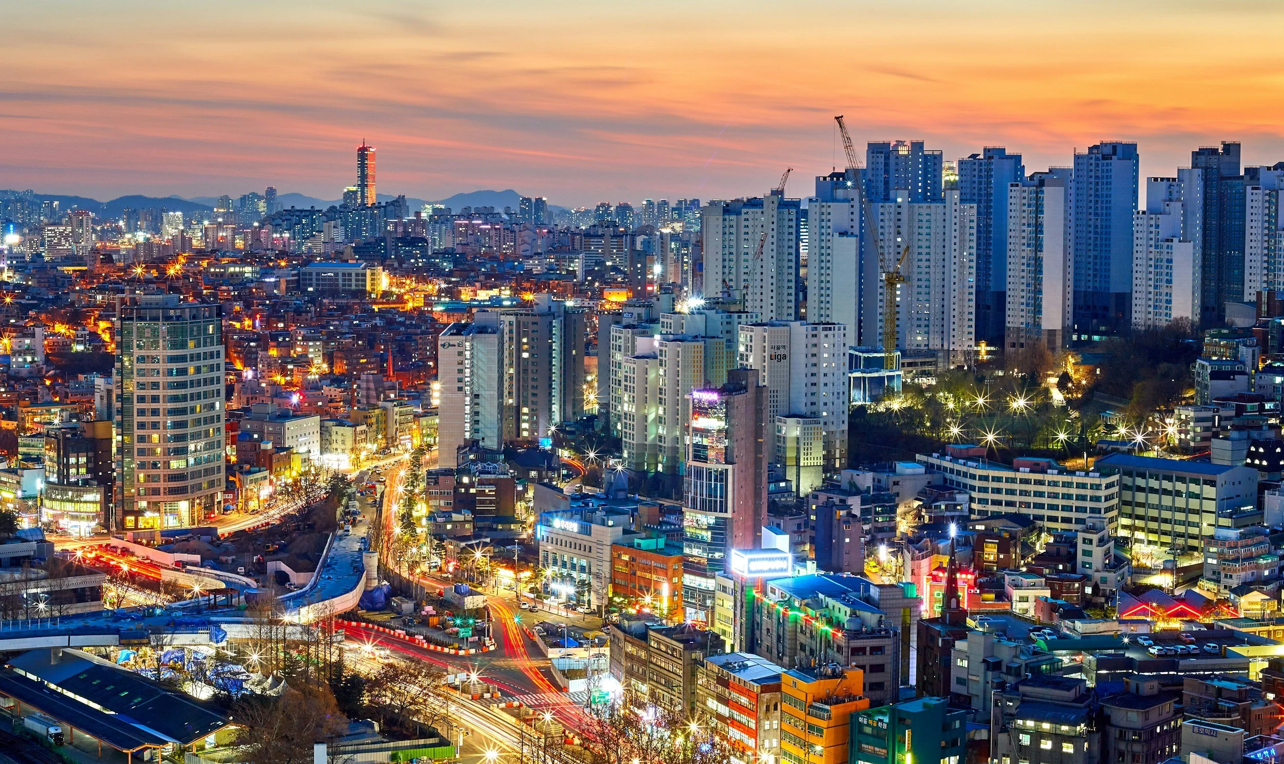 City skyline at dusk with high-rise buildings, illuminated streets, and light trails from moving vehicles.