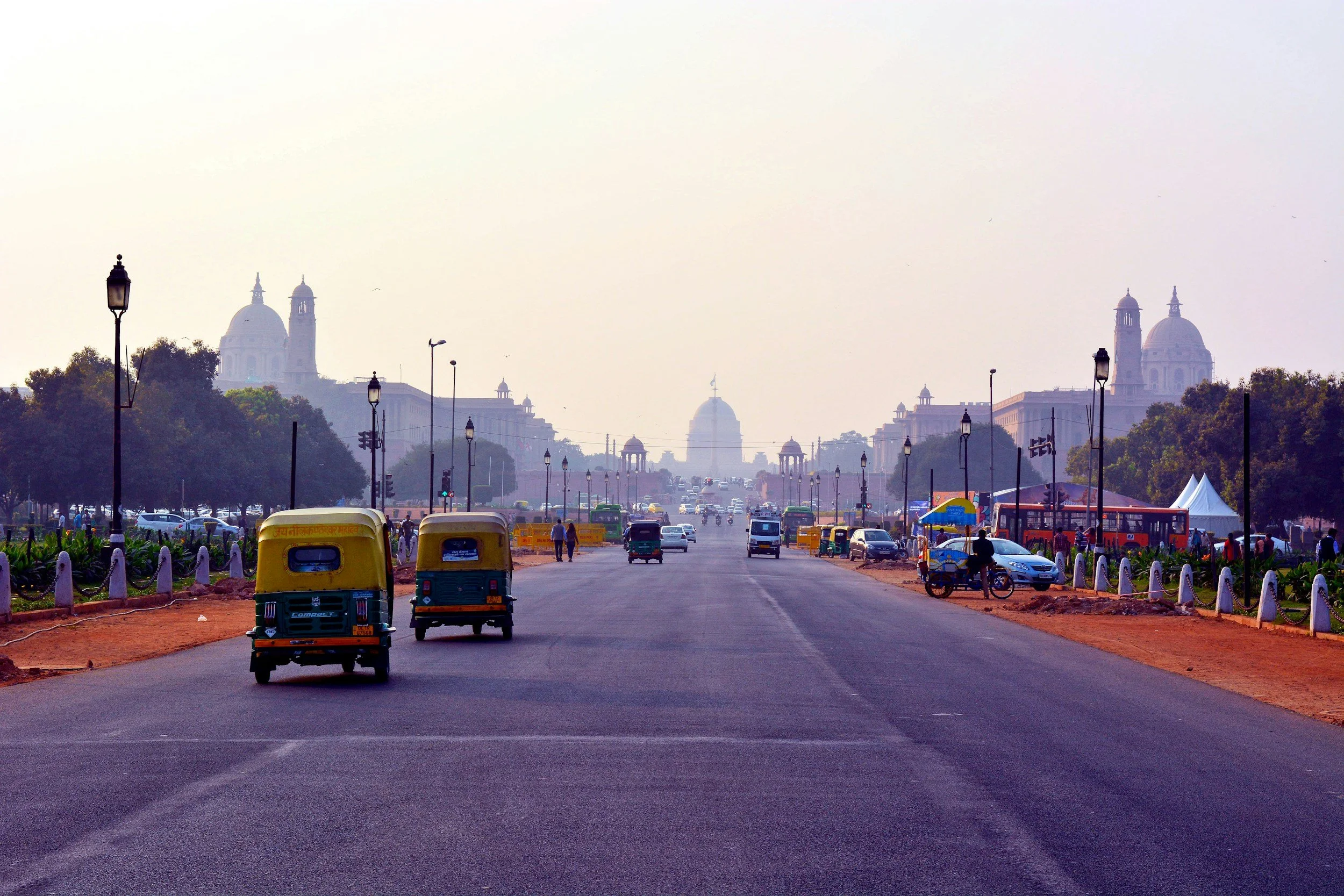 A wide street in India with auto rickshaws, cars, and buses, lined with lamp posts and trees, leading to historic buildings and domes in the distance under a hazy sky.