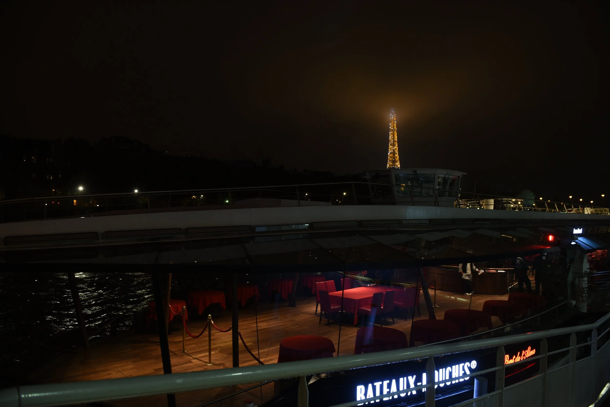 Nighttime view of a boat with a lit-up Eiffel Tower replica in the background and some illuminated signs on the boat, including 'BATEAUX-LUCHES' and 'Pont de Siene.' The boat features red-draped tables and seating, visible through windows.