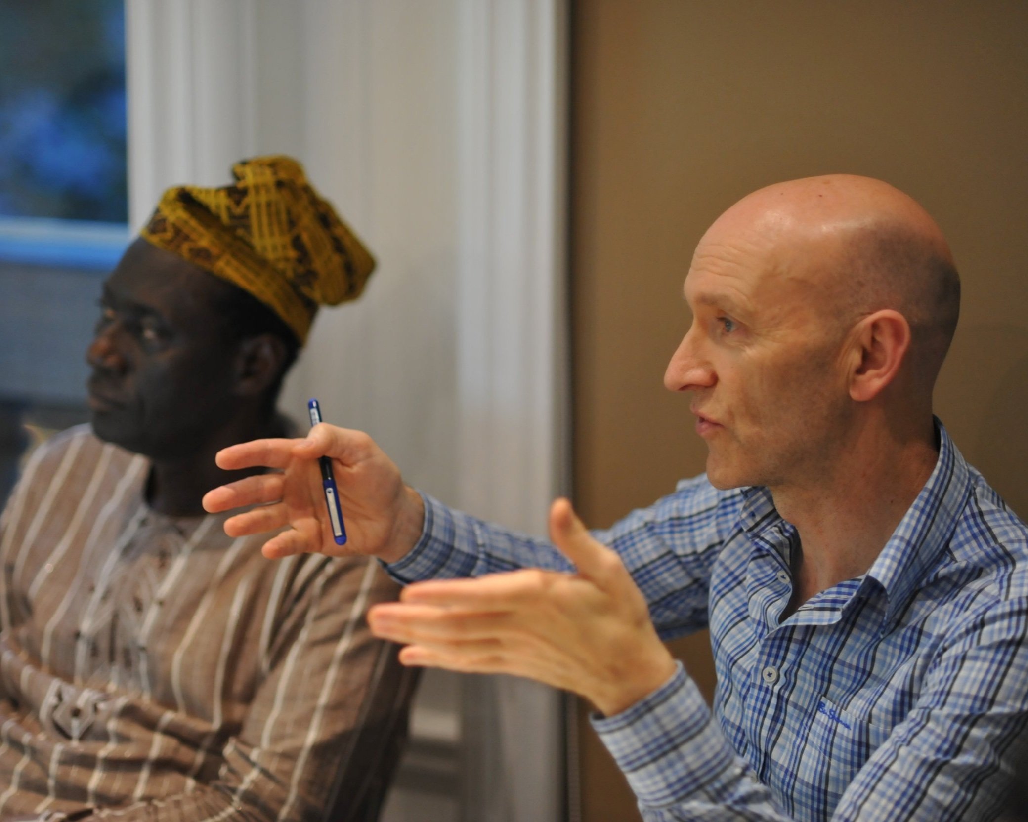 Two men are sitting at a table, engaged in a discussion. The man on the right, who is bald and wearing a blue plaid shirt, is gesturing with his hands. The man on the left, wearing a striped shirt and a colorful head wrap, is looking attentively.