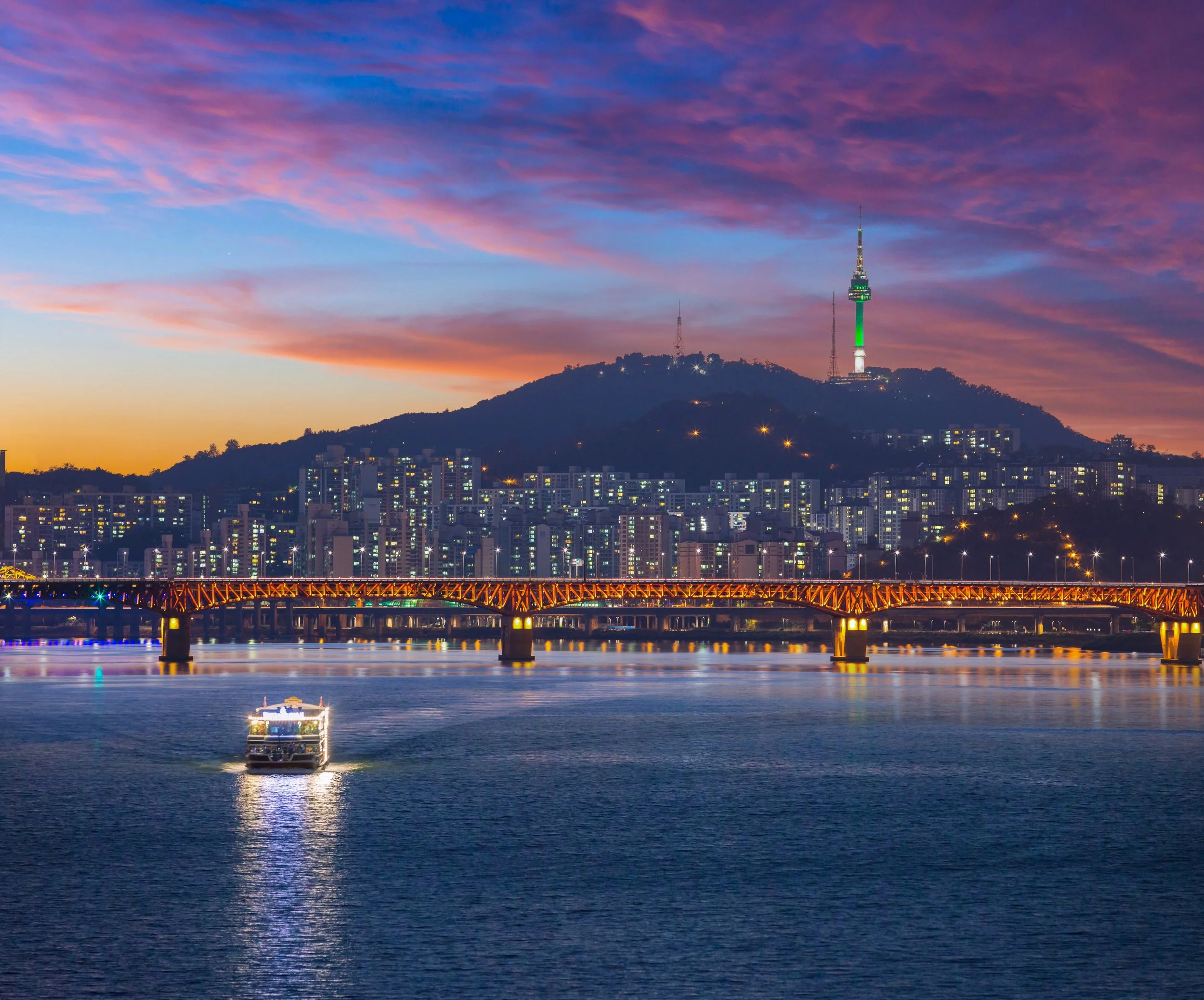 Sunset over a city with a hill topped by a tower, a bridge crossing a river, and a boat on the water.