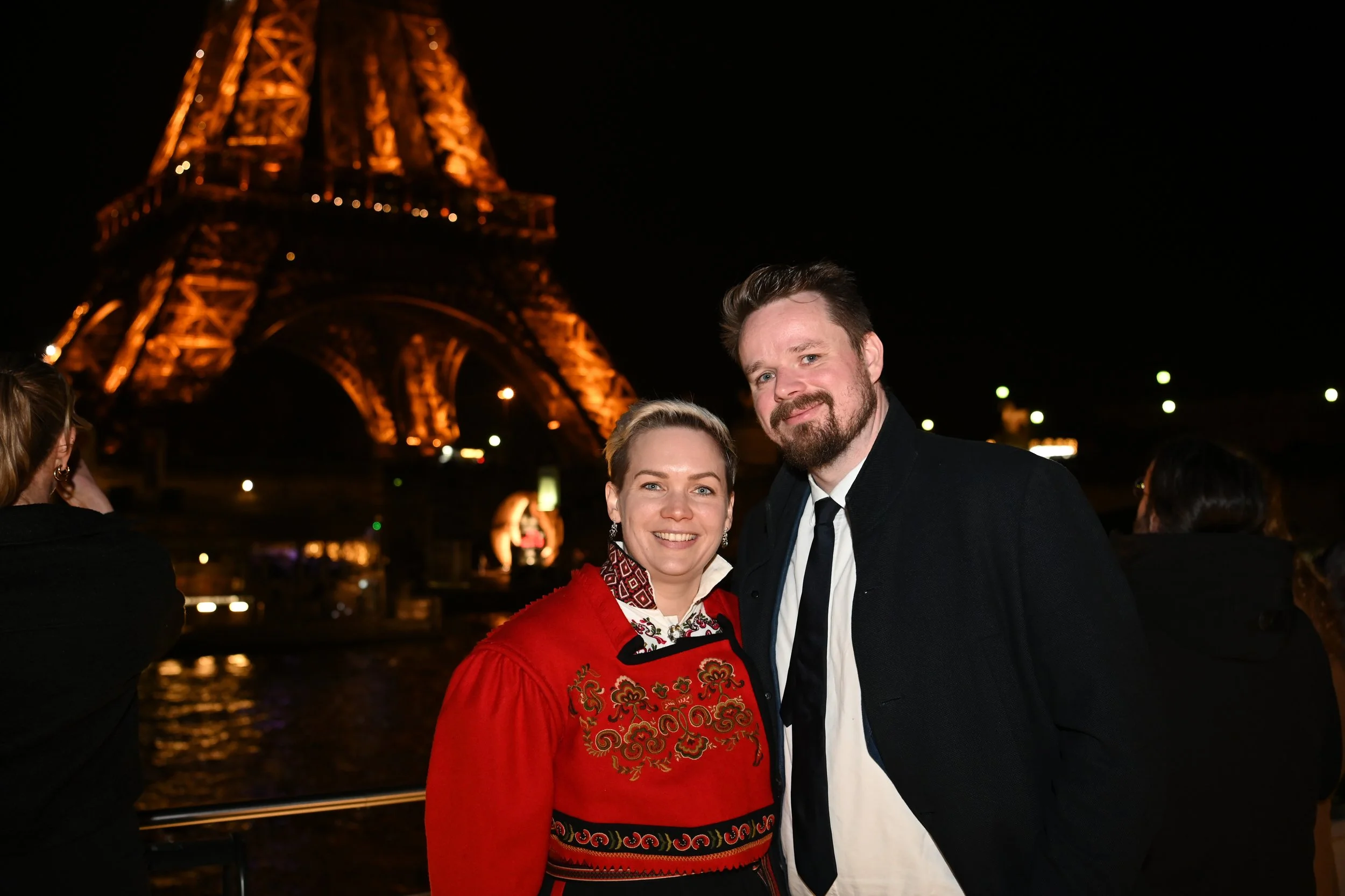 A smiling couple stands close together at night near the Eiffel Tower, which is illuminated in the background.