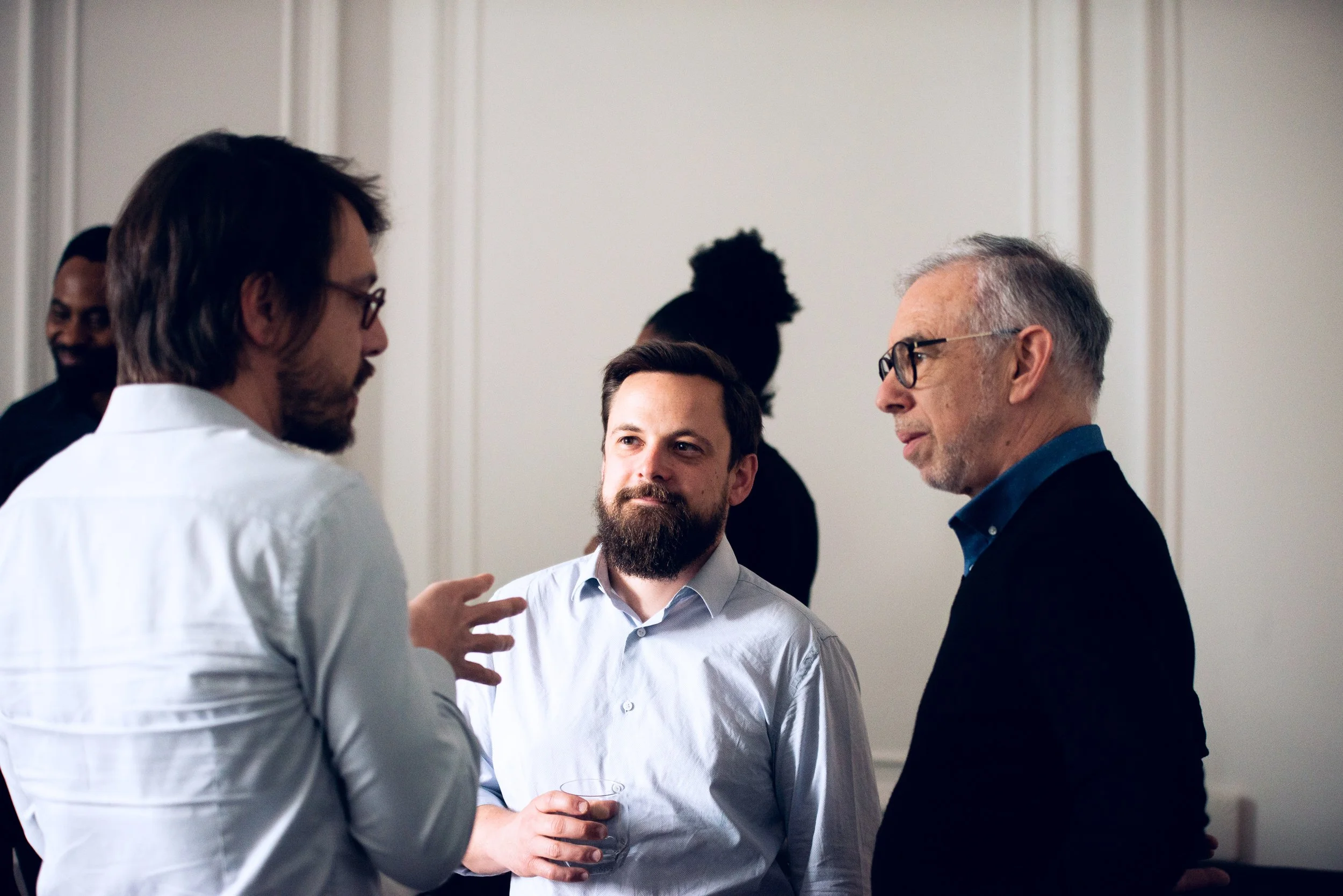Three men engaged in conversation at a social gathering. One man is gesturing with his hands, while another is holding a glass. The third man listens attentively. There are other people in the background.