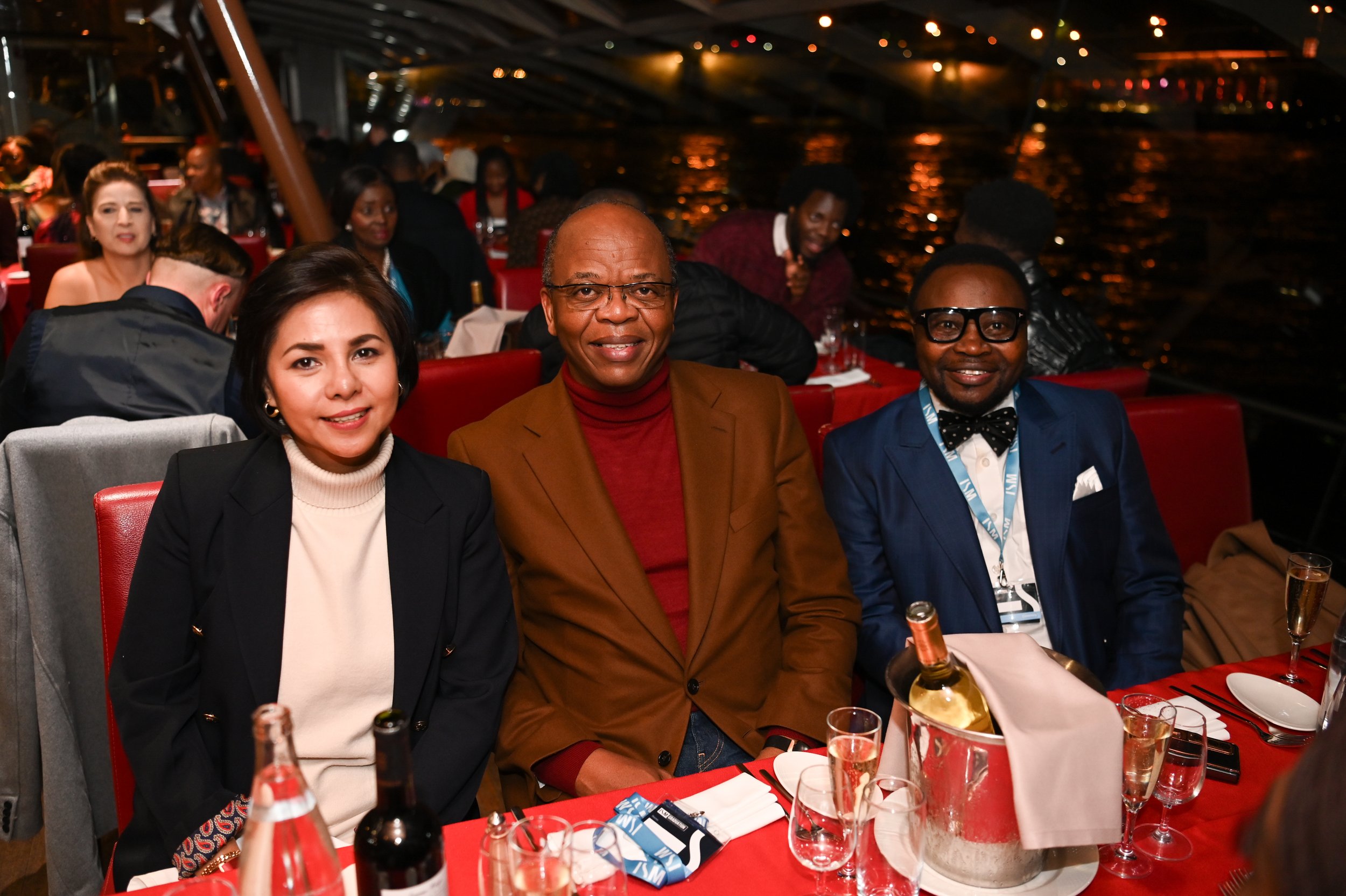 Three people sitting at a dinner table on a boat at night, with water and lights visible in the background, smiling at the camera.