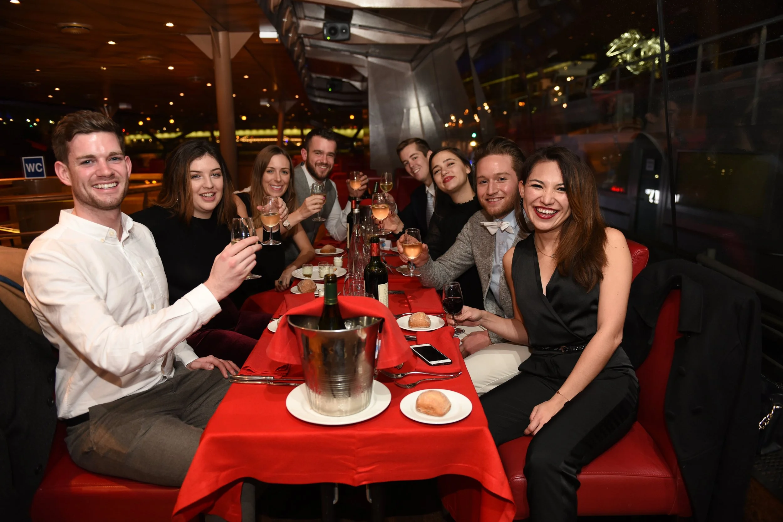 A group of ten friends gathered at a restaurant table, enjoying drinks and smiling for the camera during a night out.
