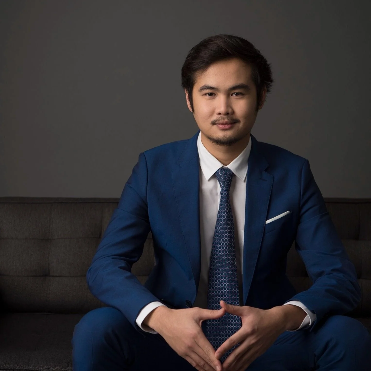 A man in a navy blue suit with a white shirt and patterned tie sitting on a dark sofa against a plain gray background, with a confident expression.