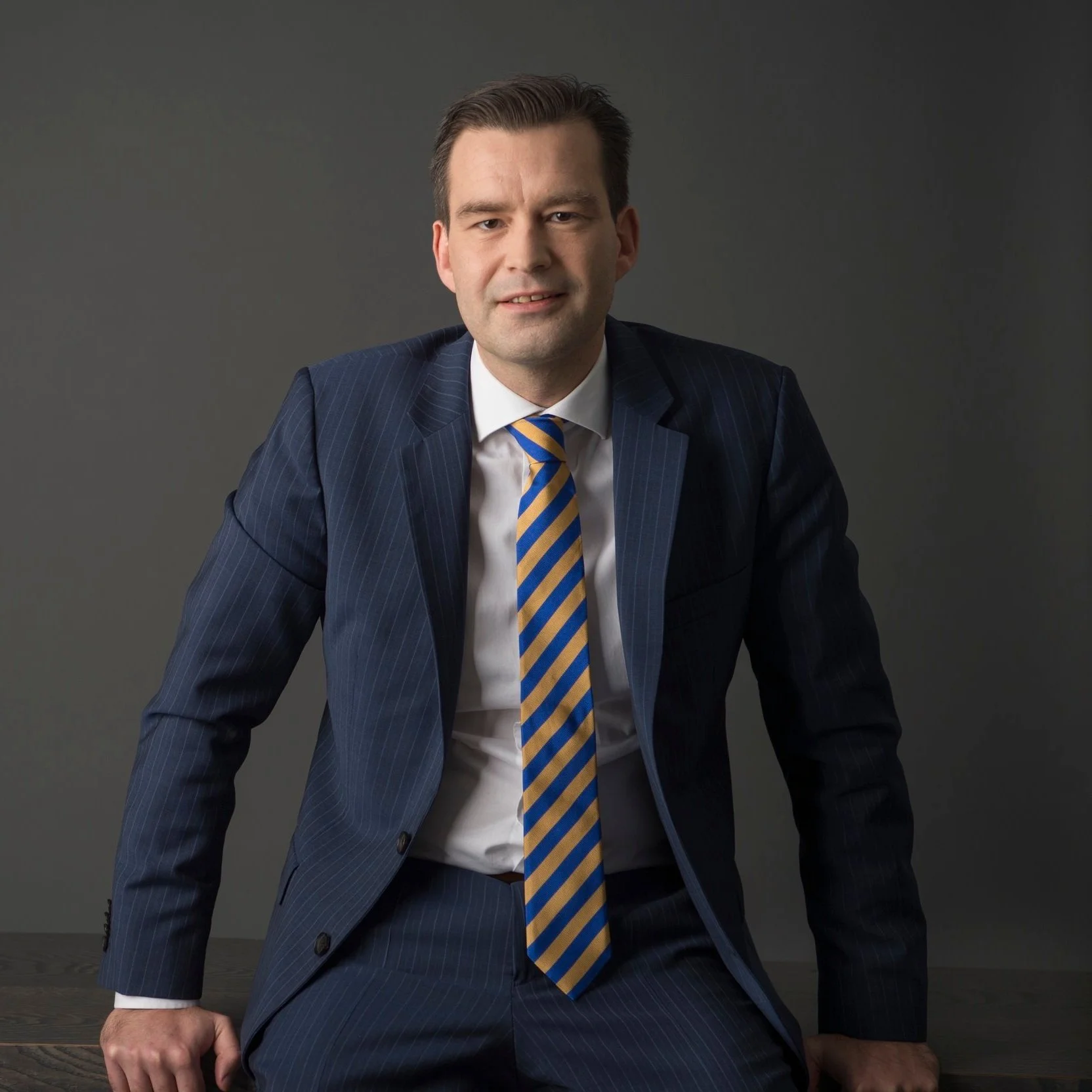 A man in a blue pinstripe suit, white shirt, and striped yellow and blue tie, sitting at a dark wooden table against a plain gray background.