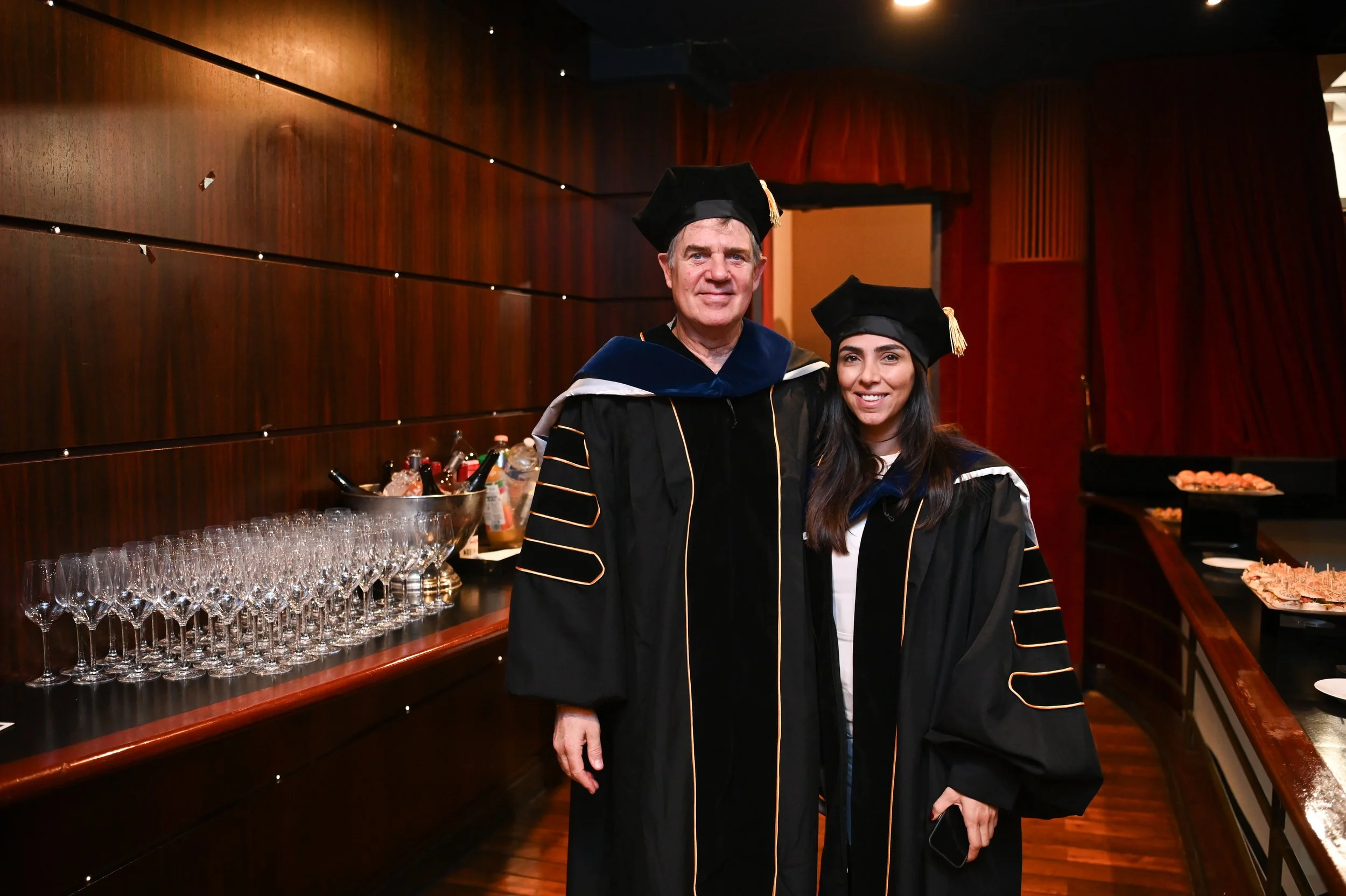 Two people in academic regalia at a graduation ceremony, standing in a room with wooden paneling and a table with glasses and drinks in the background.