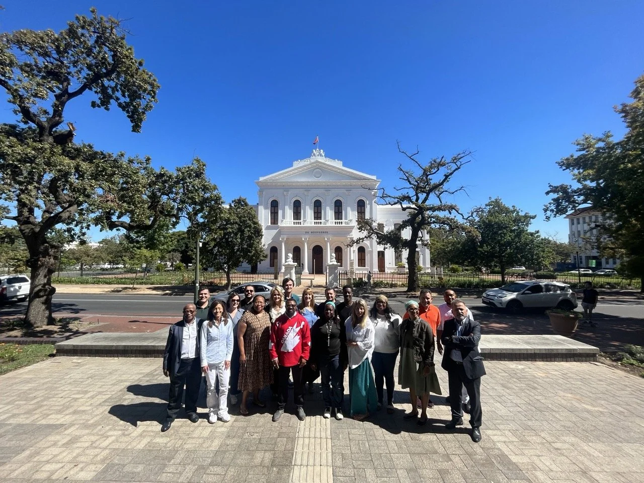 Group of people standing on sidewalk in front of the Alabama State Capitol building, with trees and cars visible in the background on a bright, sunny day.