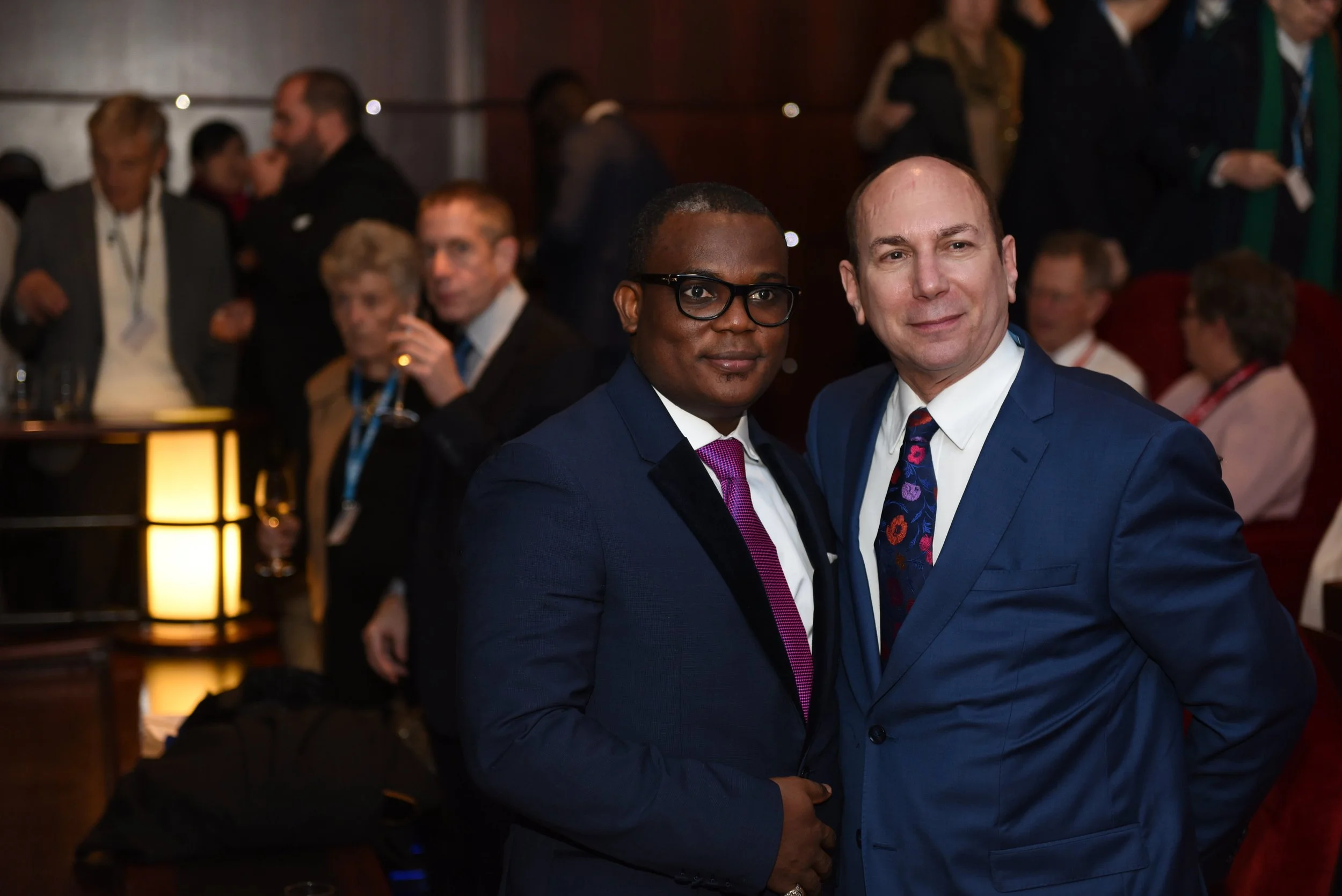 Two men in suits posing together at a professional event or conference, with other attendees mingling in the background.