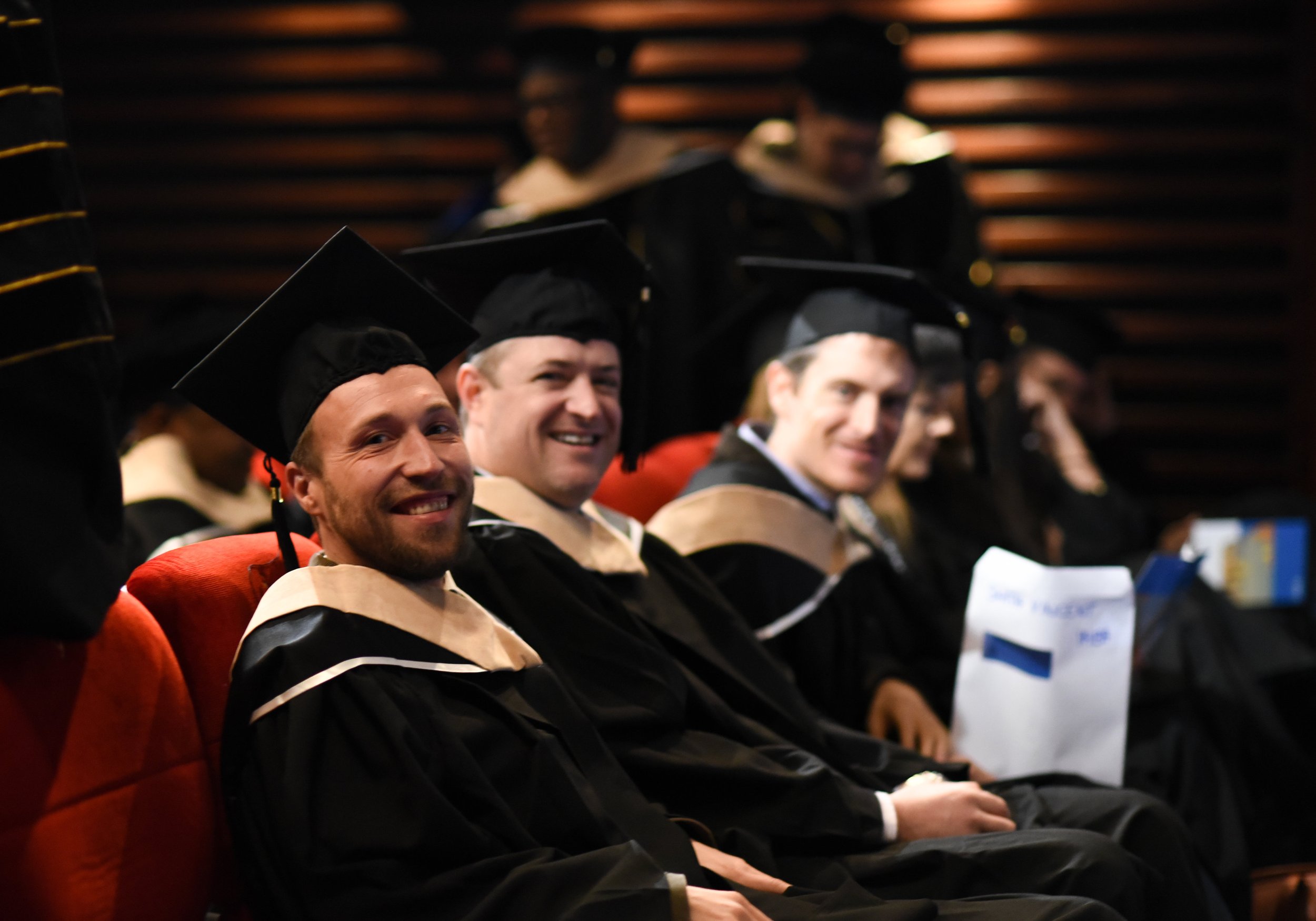 Graduates in black caps and gowns with beige stoles, seated and smiling during a graduation ceremony.
