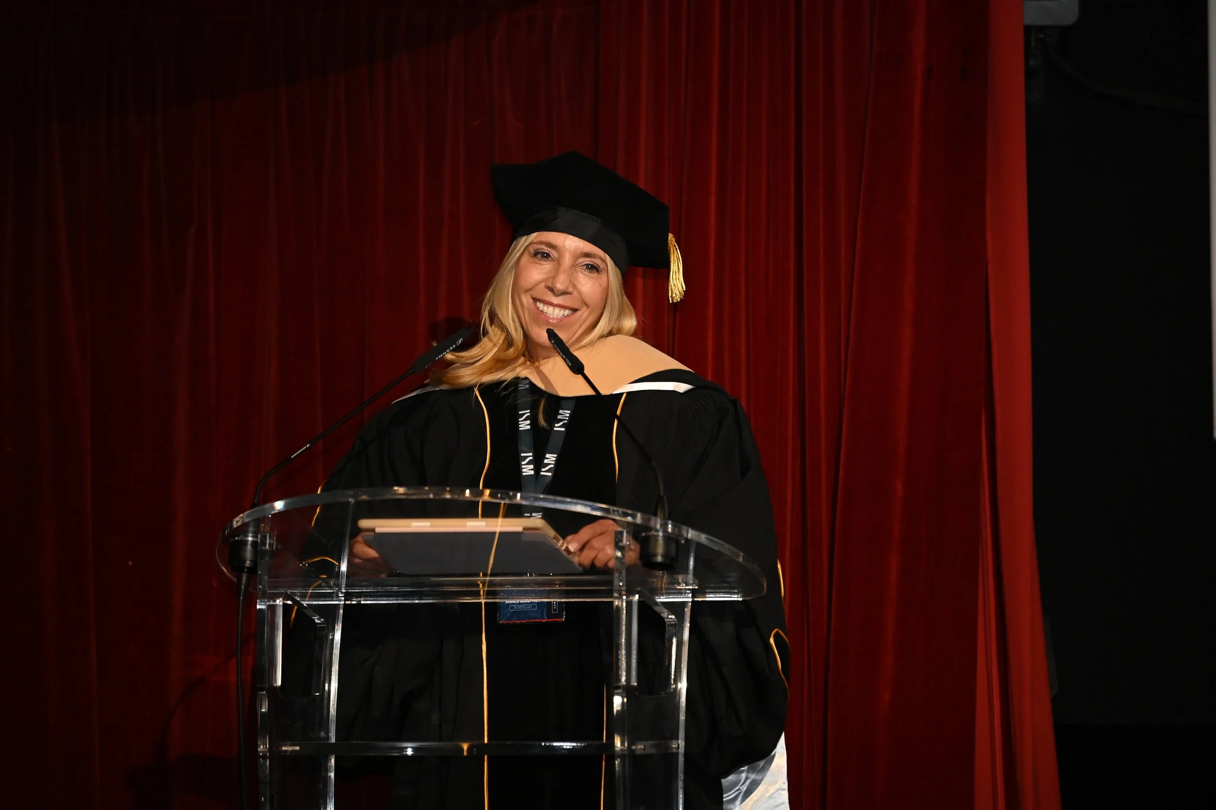 Woman in academic regalia smiling at a podium during a graduation ceremony.