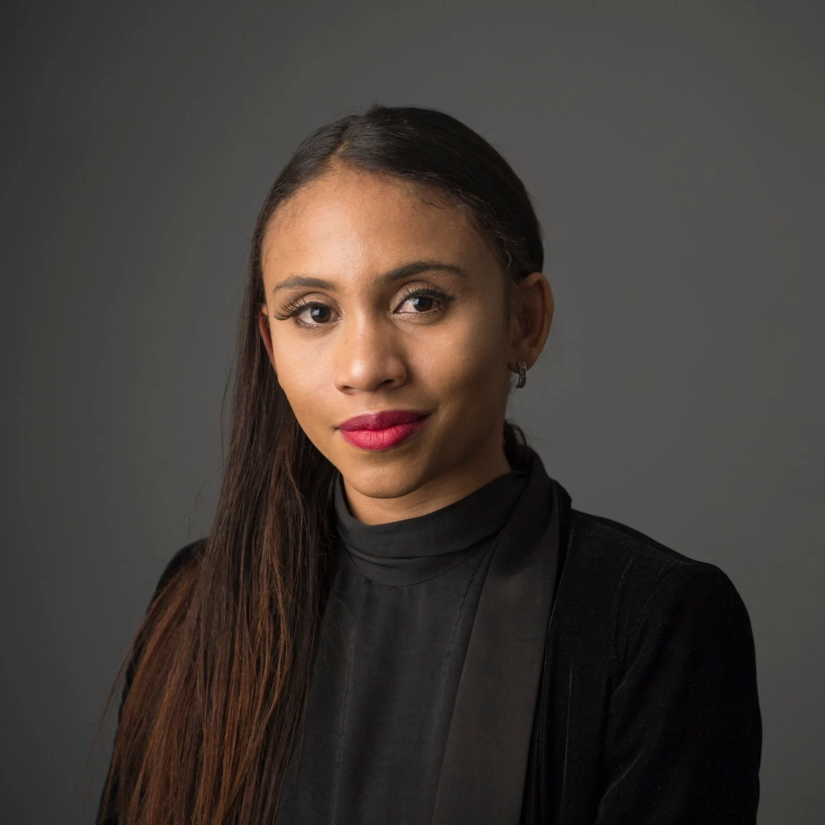 Portrait of a woman with long dark hair, wearing a black top, against a gray background.