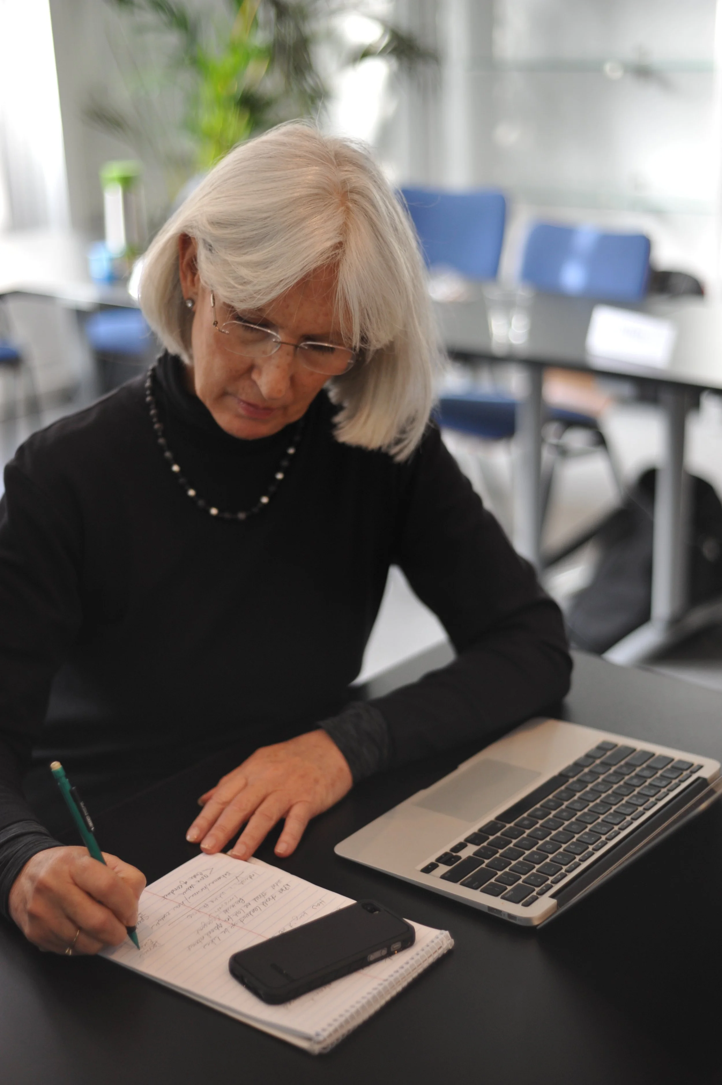 An older woman with gray hair and glasses, wearing a black top and a pearl necklace, is sitting at a table in a conference room. She is writing notes on a notepad while using a laptop, with a smartphone placed on the notepad. The room has blue chairs, large windows, and some potted plants in the background.