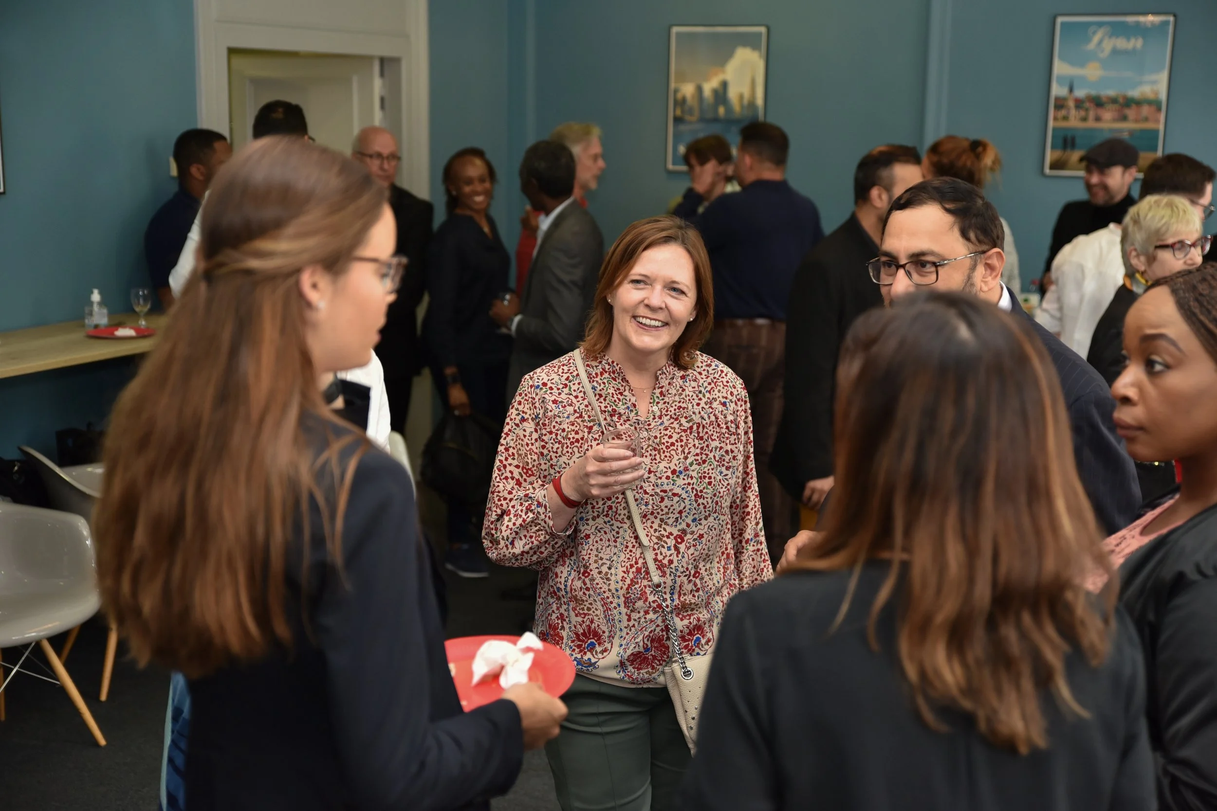 People socializing at a networking event, mingling and smiling in a room with framed artwork on teal walls.