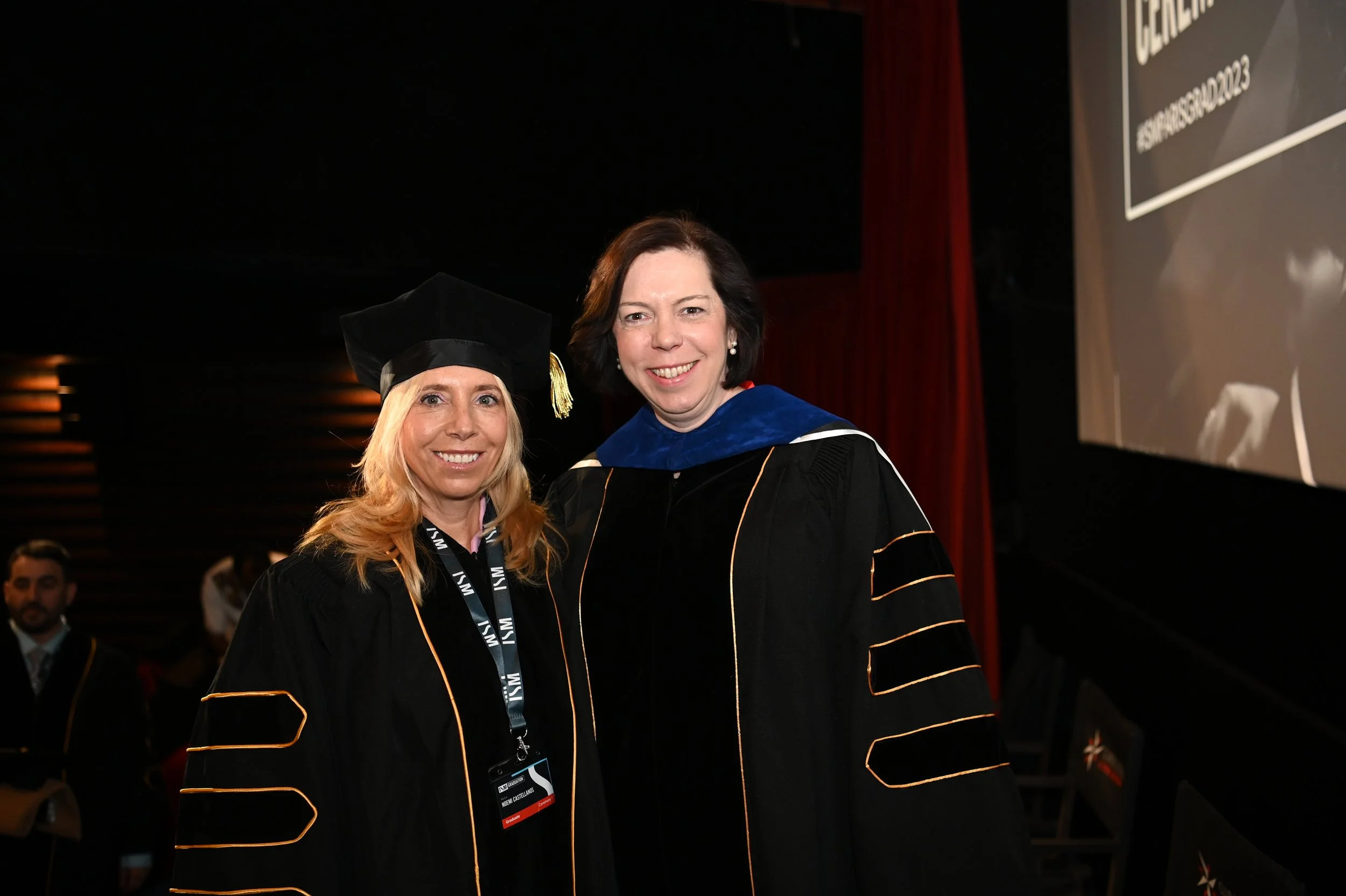 Two women in academic regalia at a graduation ceremony, smiling and standing side by side.