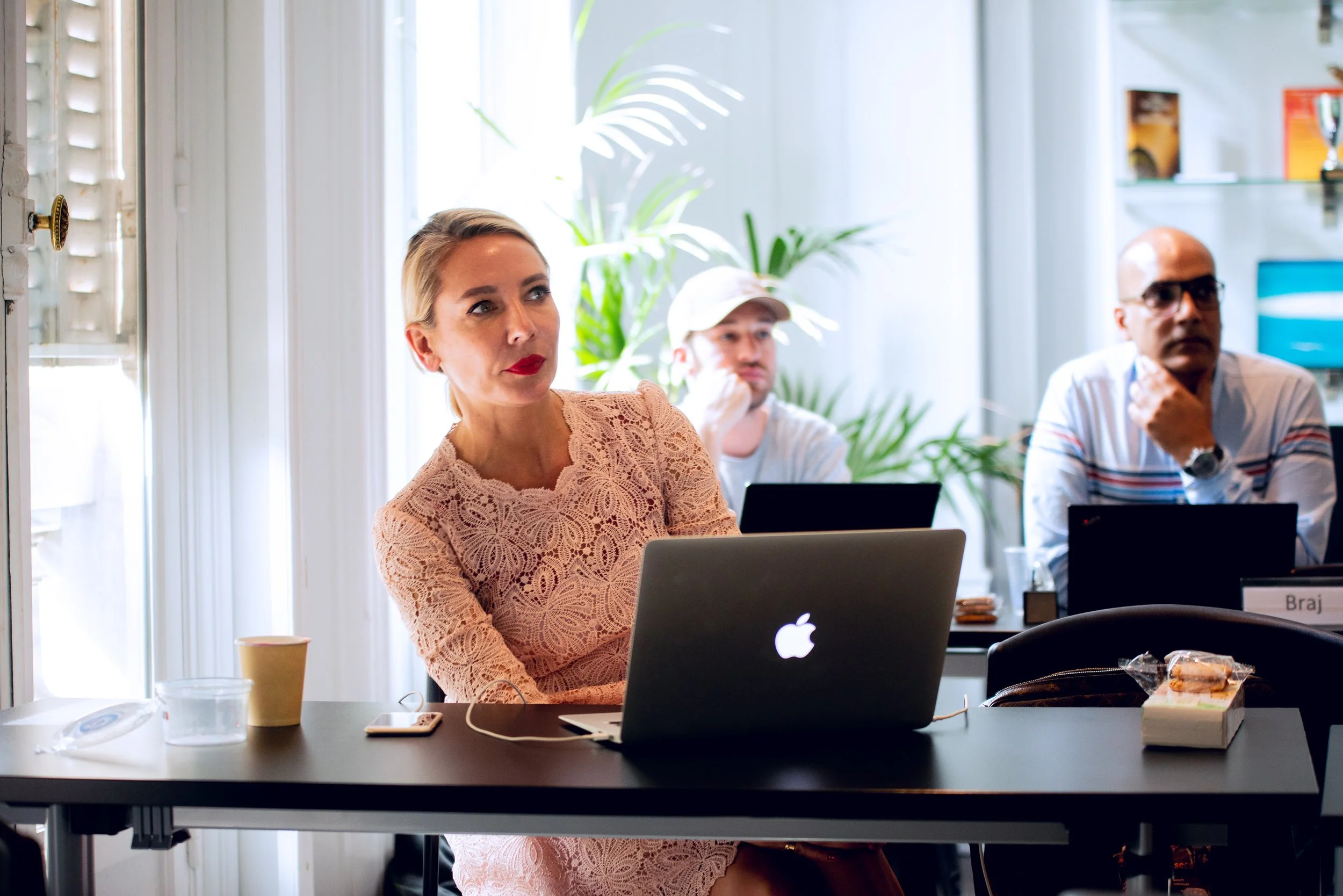 A woman with blonde hair wearing red lipstick and a lace beige top sitting at a desk with a silver Apple MacBook laptop, a white smartphone, a paper cup, and a glass of water. Behind her, a man in a white cap and gray T-shirt and another man in glass