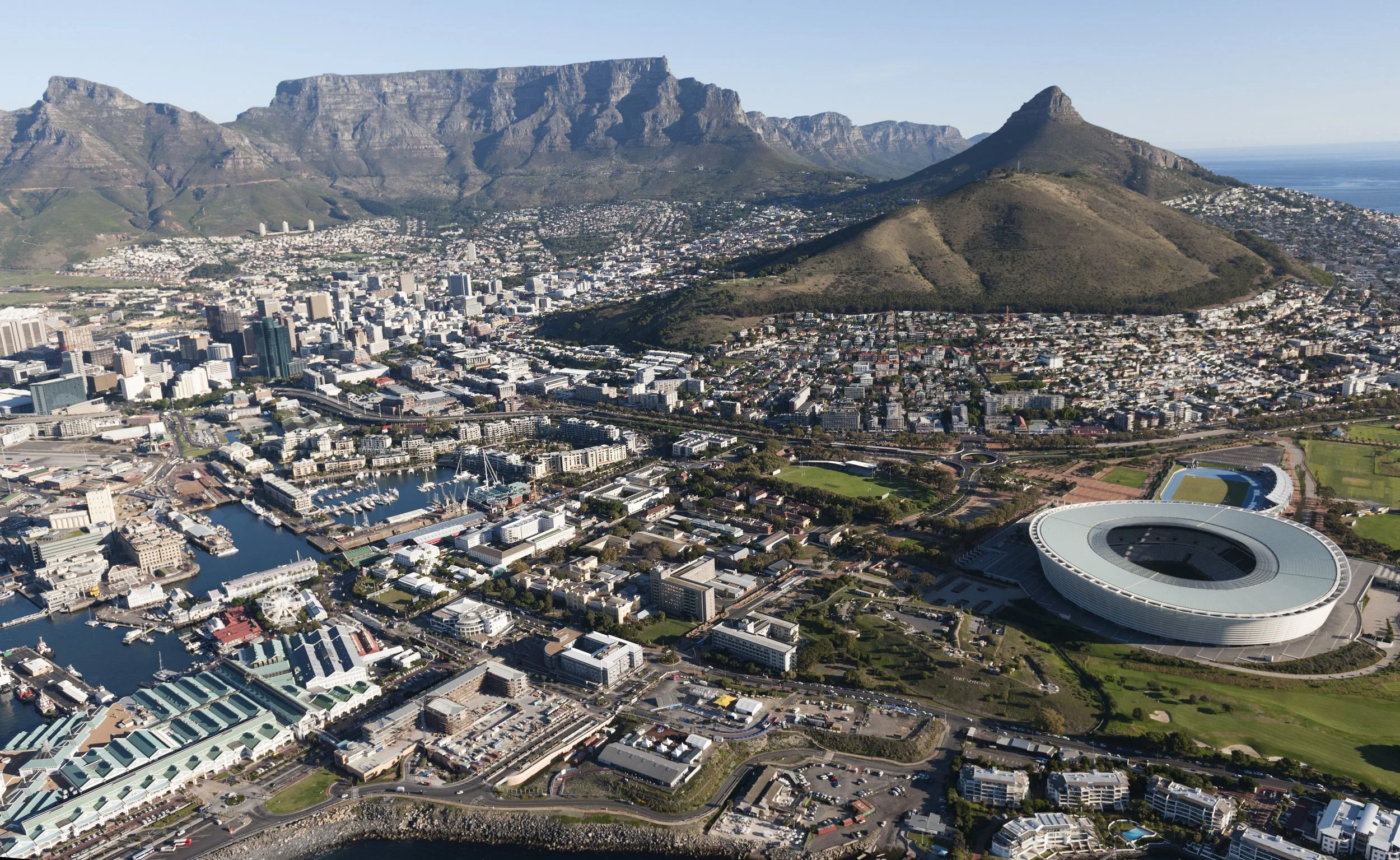 Aerial view of Cape Town, South Africa, showing the city skyline, harbor, Table Mountain, and Lion's Head mountain with blue sky overhead.
