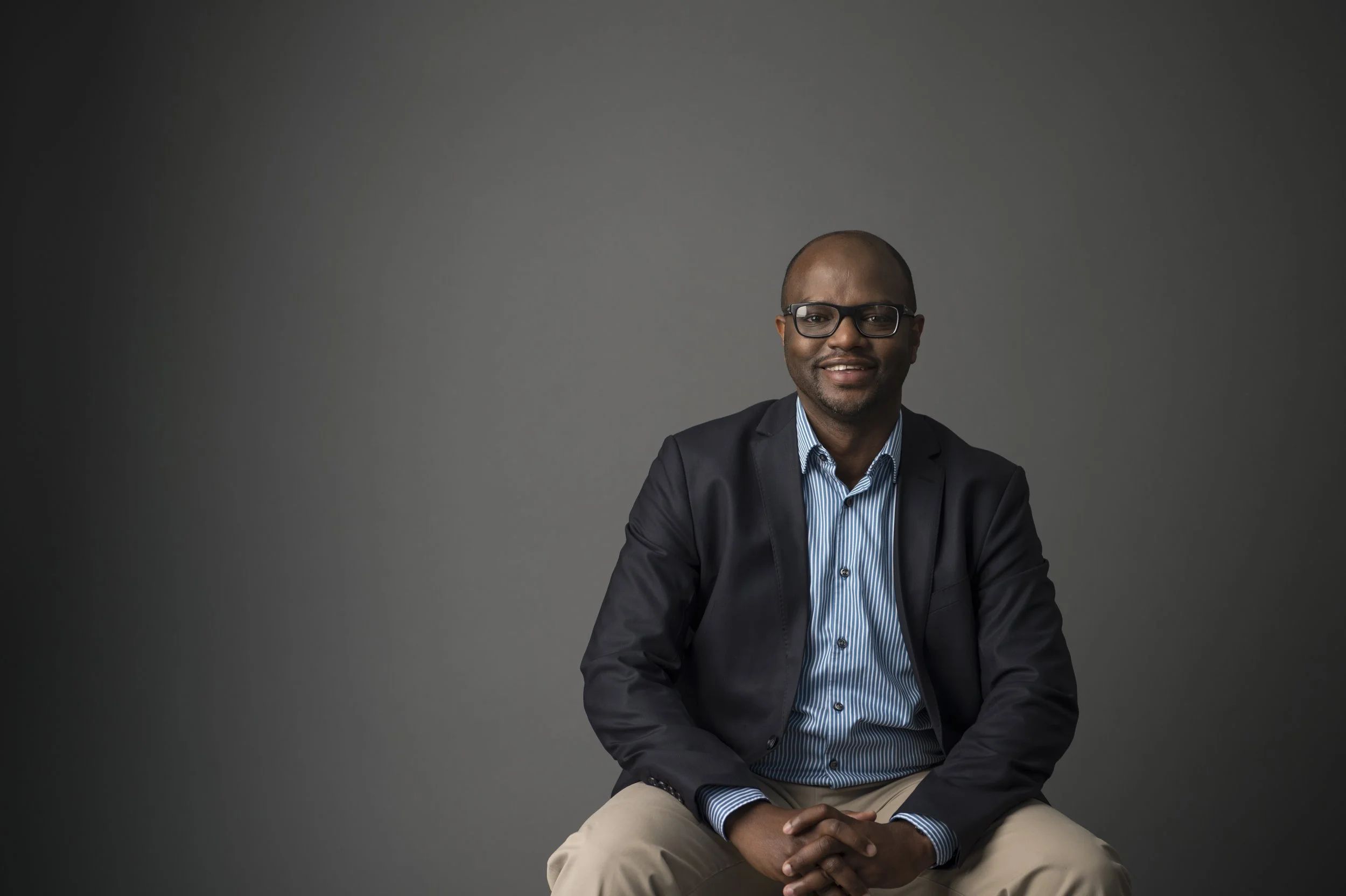 A man sitting with hands clasped, wearing glasses, a black blazer, blue striped shirt, and beige pants, smiling against a dark grey background.