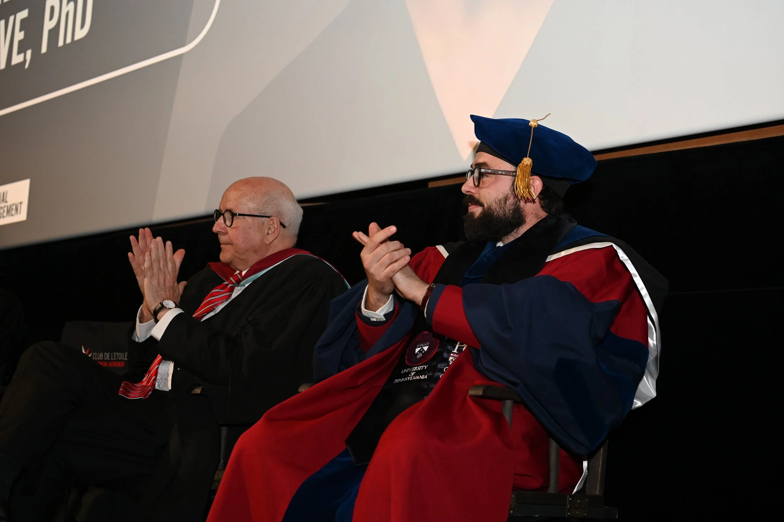 Two men sitting on a stage during a graduation ceremony, wearing academic regalia. The man on the right is dressed in a blue and red gown with a matching cap with gold tassels, and has a beard and glasses. The man on the left has white hair, glasses,