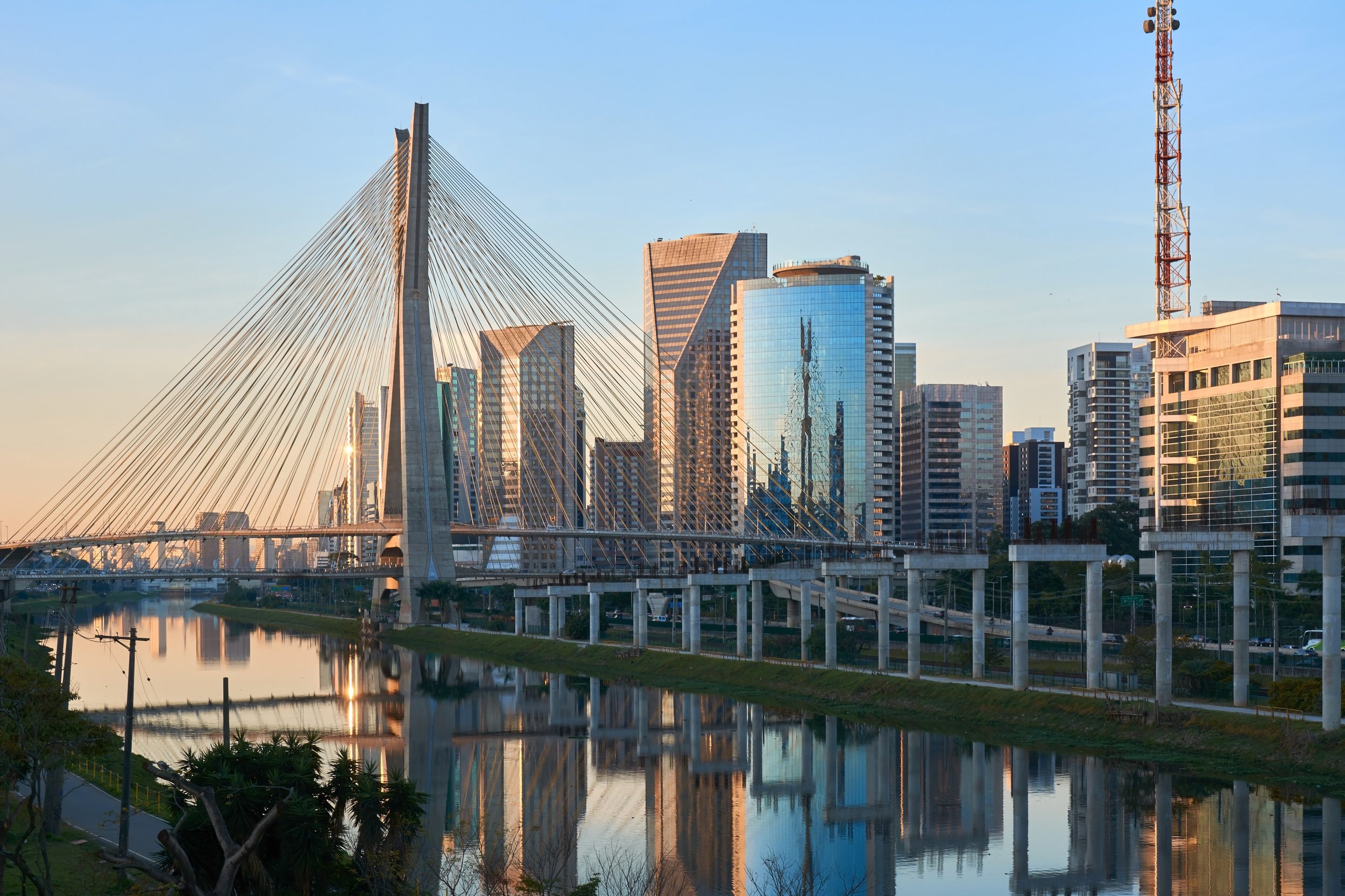 City skyline with modern high-rise buildings and a cable-stayed bridge over a river at sunset.
