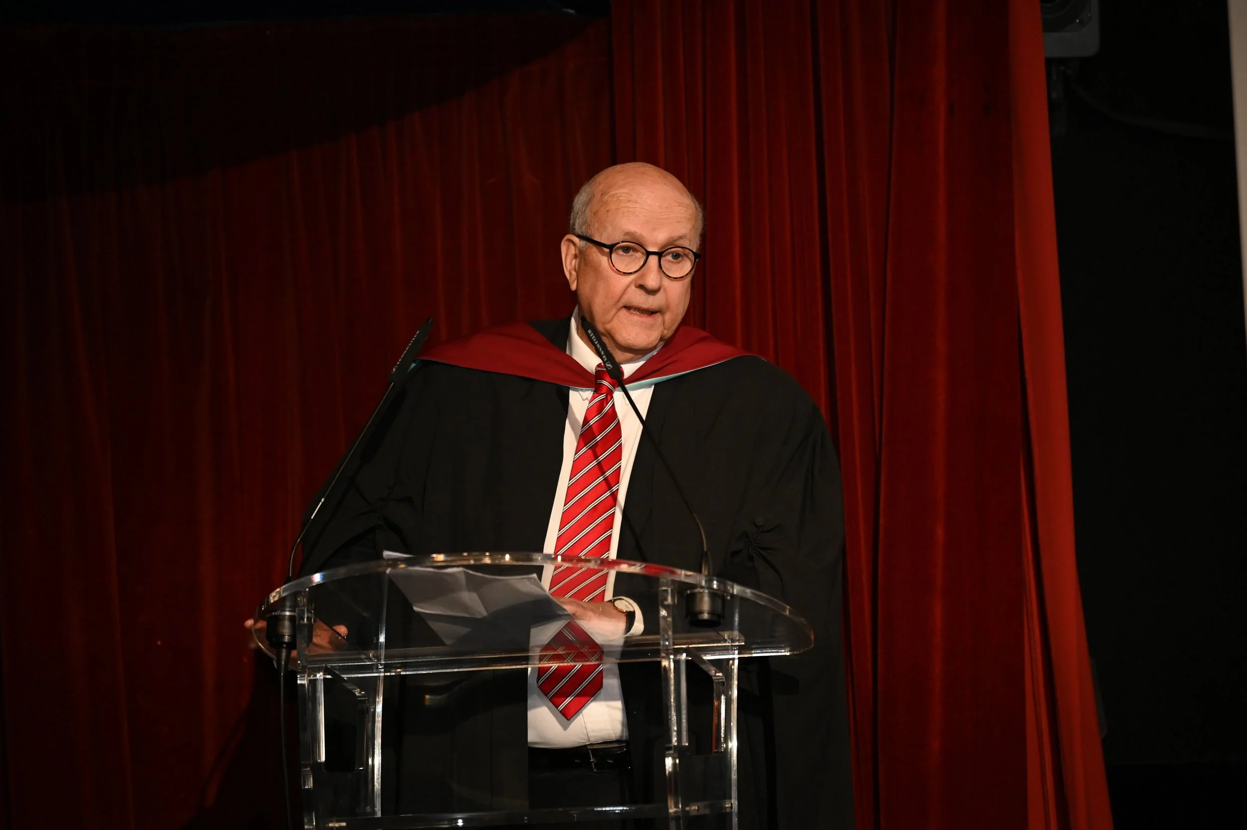 An elderly man in academic regalia, including a black gown and red mortarboard, stands at a clear podium with a red curtain backdrop, delivering a speech or lecture.