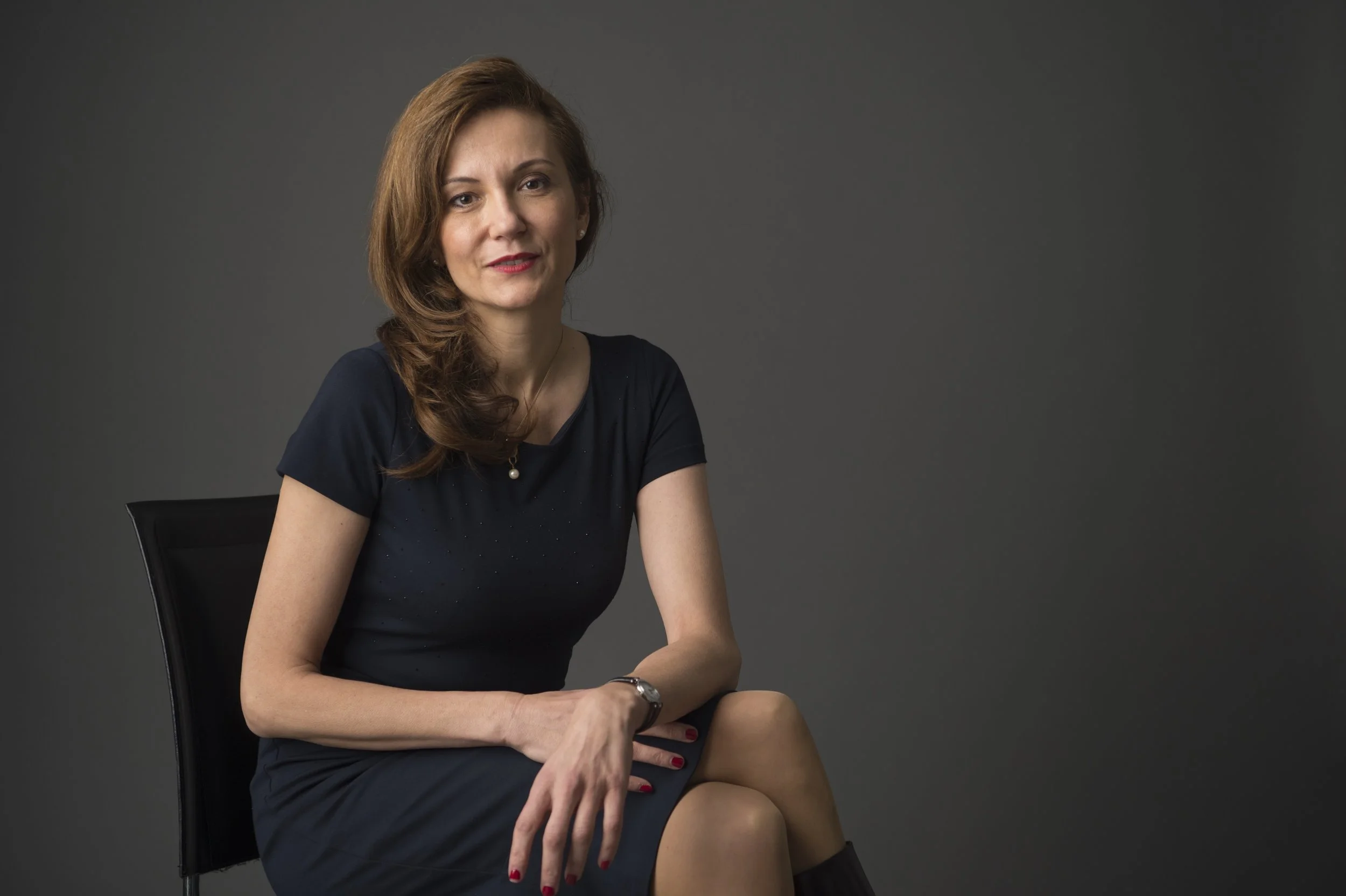 A woman with long brown hair sitting on a black chair against a gray background, wearing a navy blue dress with a pearl necklace and a watch on her left wrist.