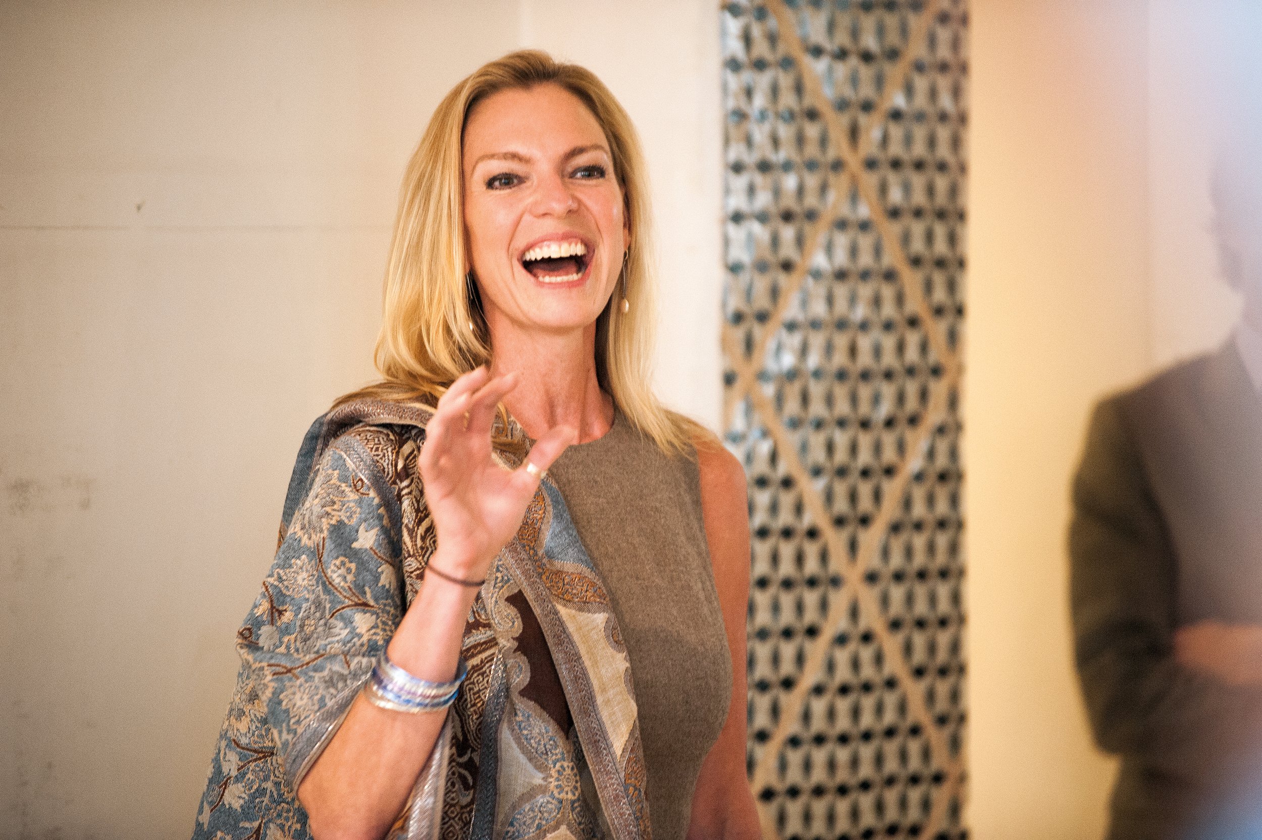 A woman with blonde hair, wearing a patterned jacket and a sleeveless brown top, is smiling and gesturing with her hand in a lively setting.