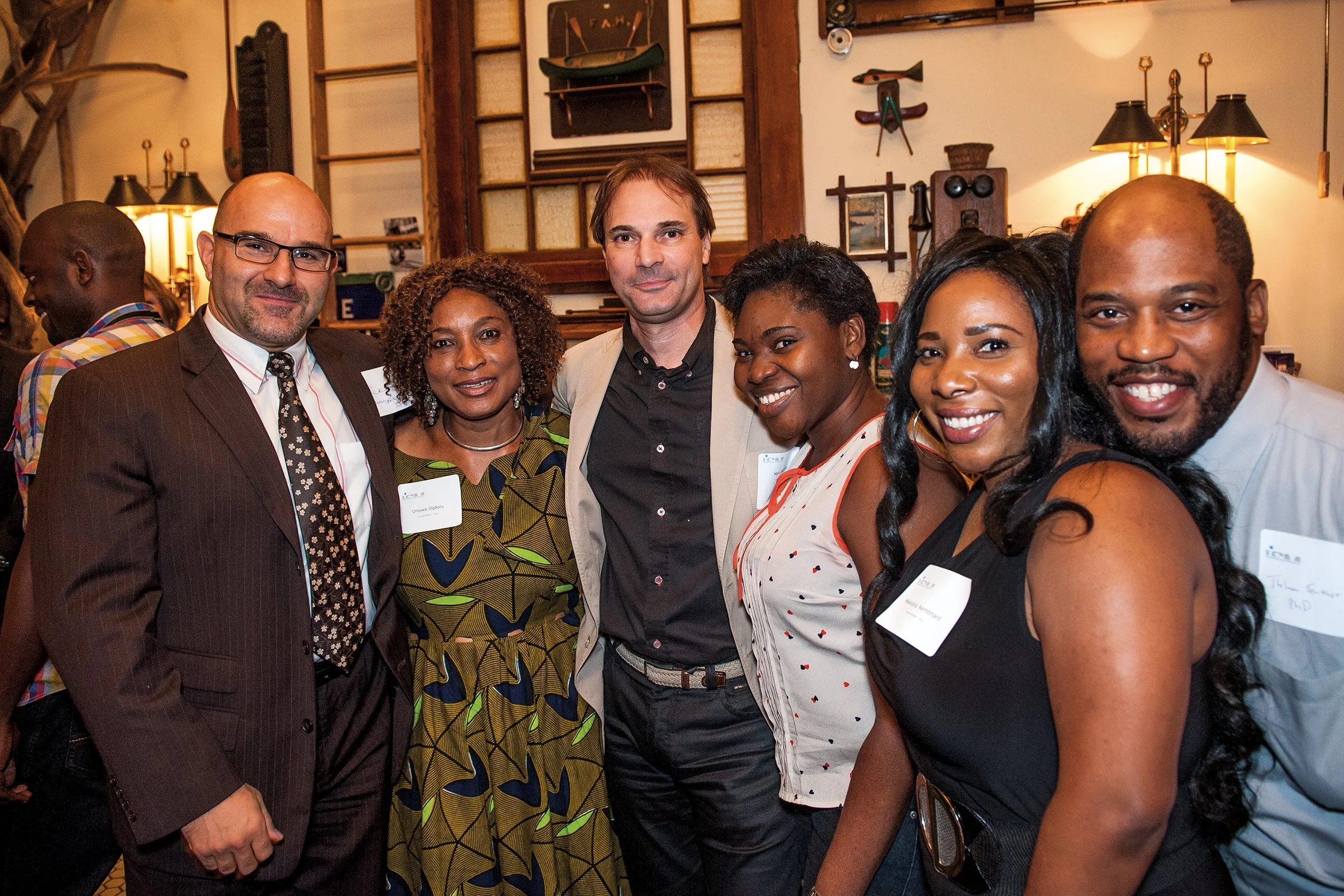 Group of six diverse adults smiling and standing together at a social event, indoors, with artwork and warm lighting in the background.