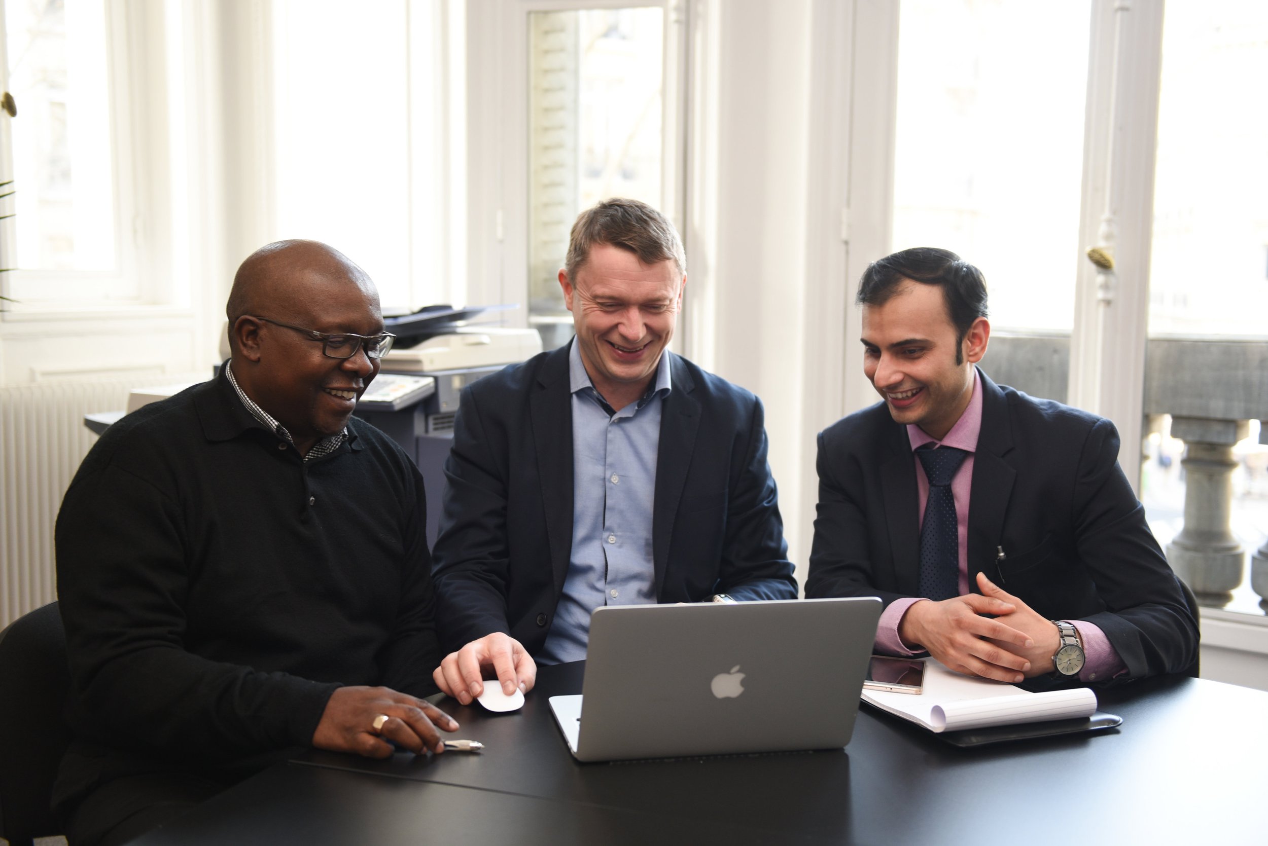 Three men sitting at a table in a bright office, looking at a laptop and sharing a smile.