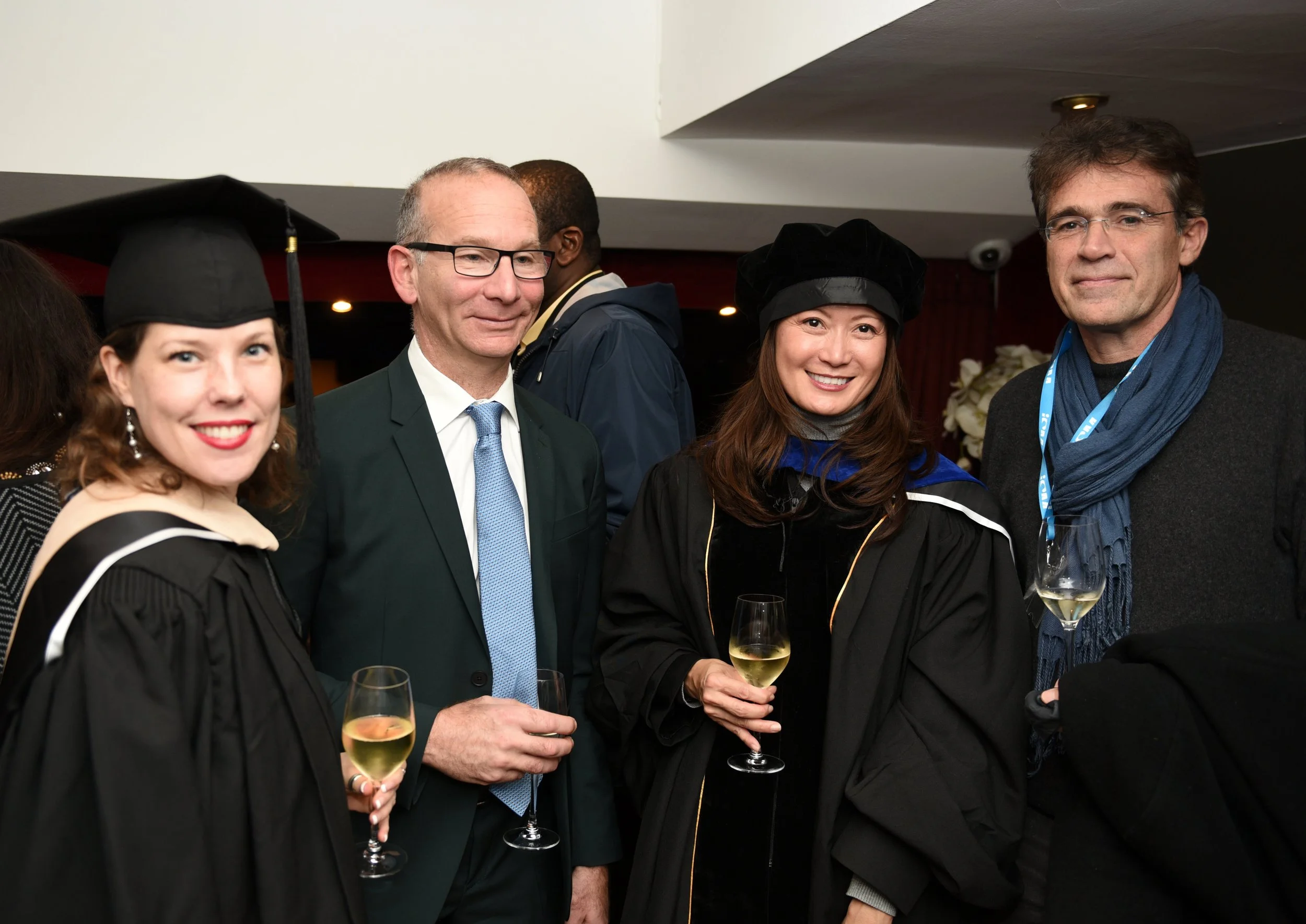 Group of four people celebrating at a graduation ceremony, two women in caps and gowns holding glasses of wine, and two men in formal wear also holding glasses of wine.