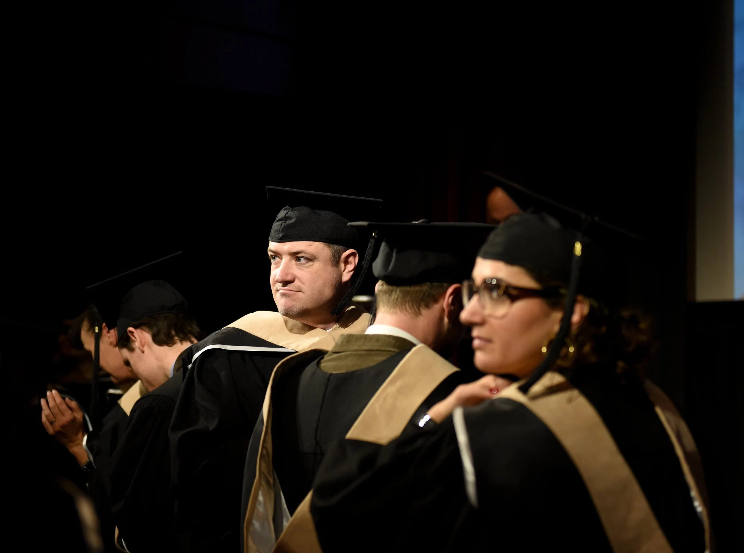 Graduates in caps and gowns sitting during a ceremony, some praying and others looking ahead.