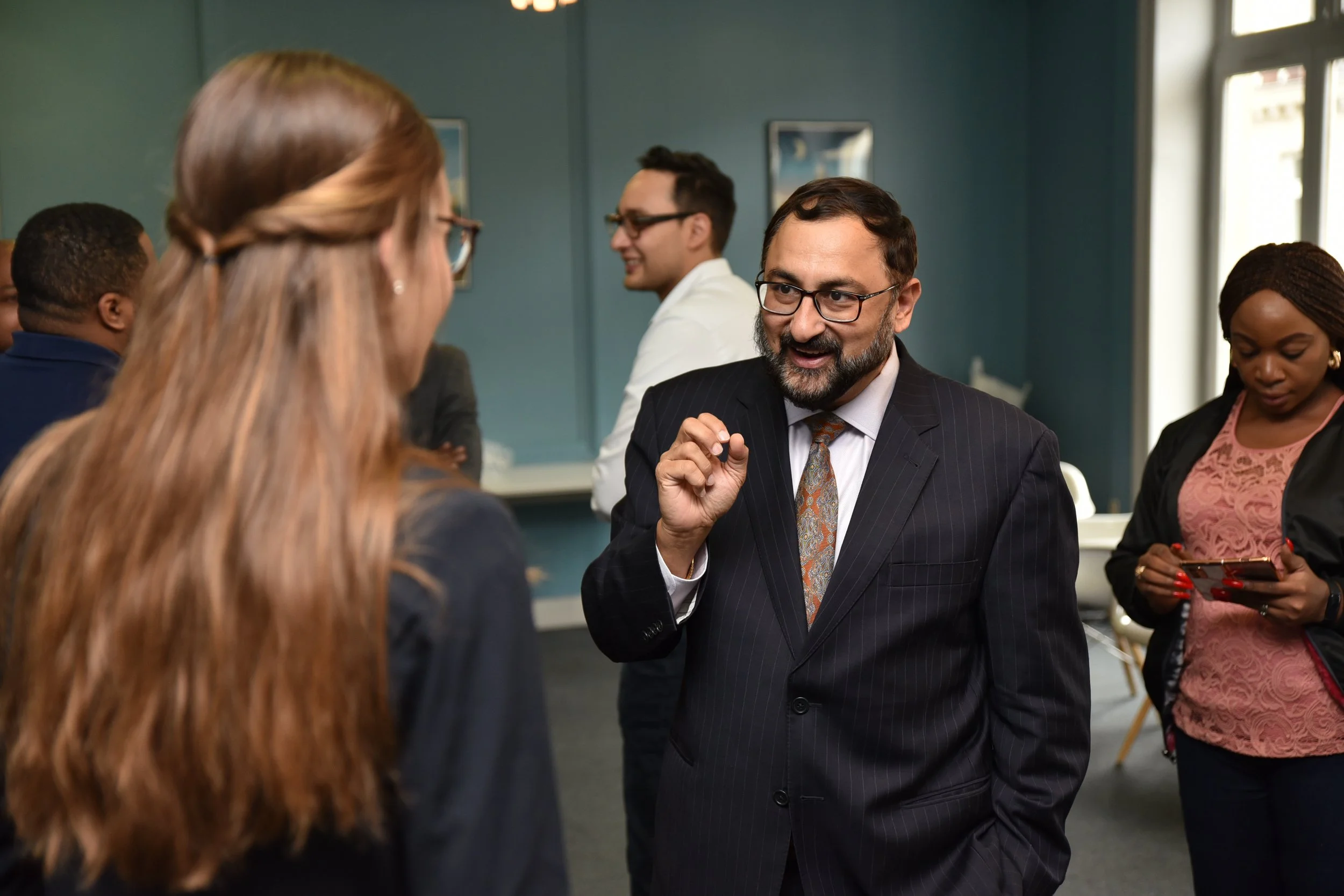 A man with glasses, a beard, and a dark pinstripe suit is talking to a woman with long, wavy, light brown hair. They are in a professional setting with other people, some of whom are engaged with their phones or chatting.